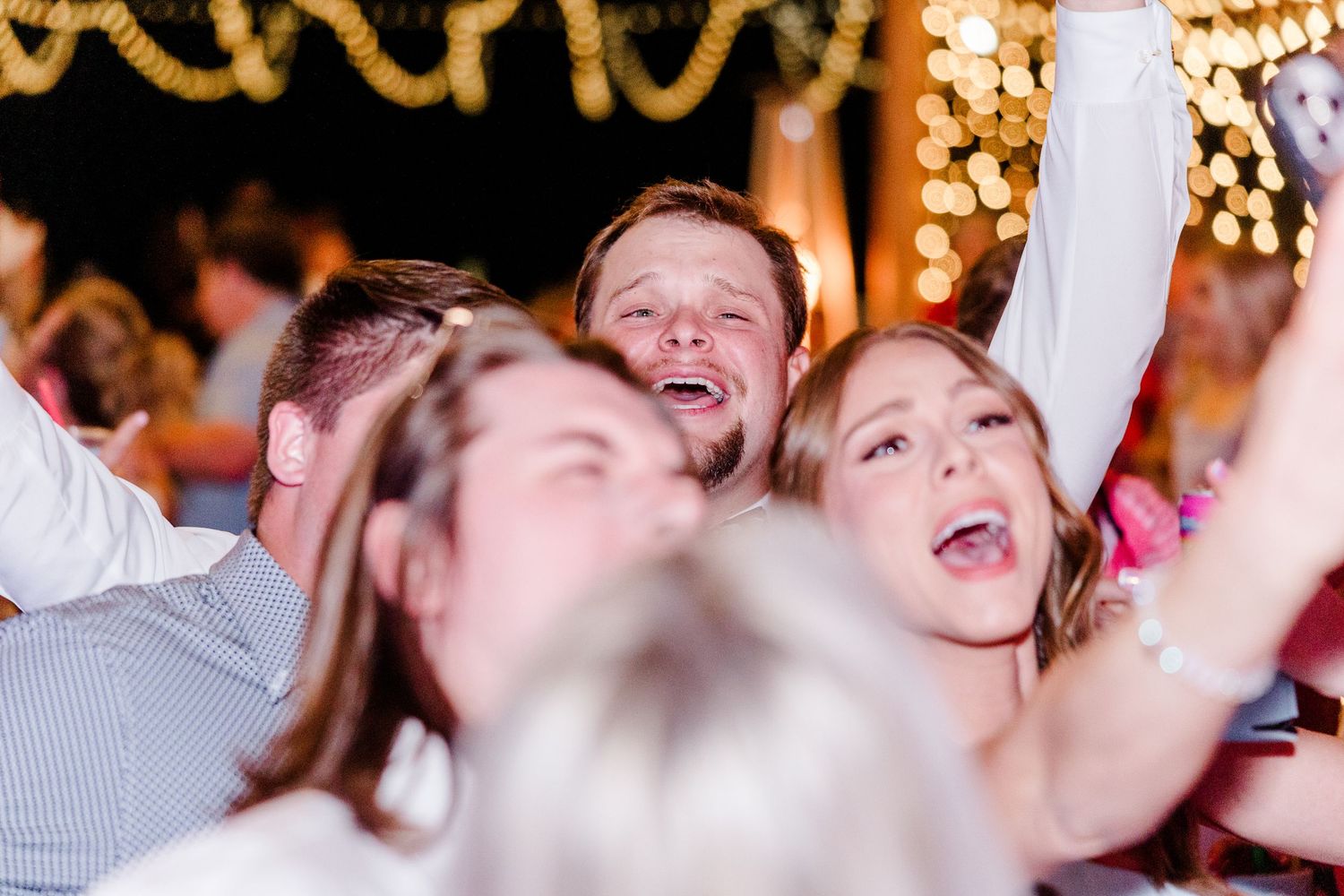 Wedding reception crowd dances enthusiastically under warm lighting in a rustic venue.