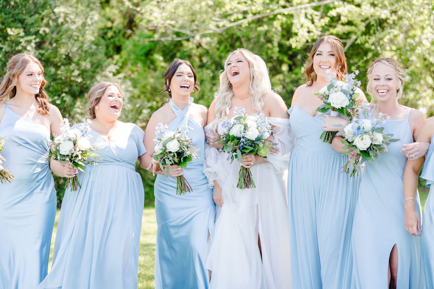 Row of bridesmaids in light blue dresses holding white and blue bouquets outdoors.