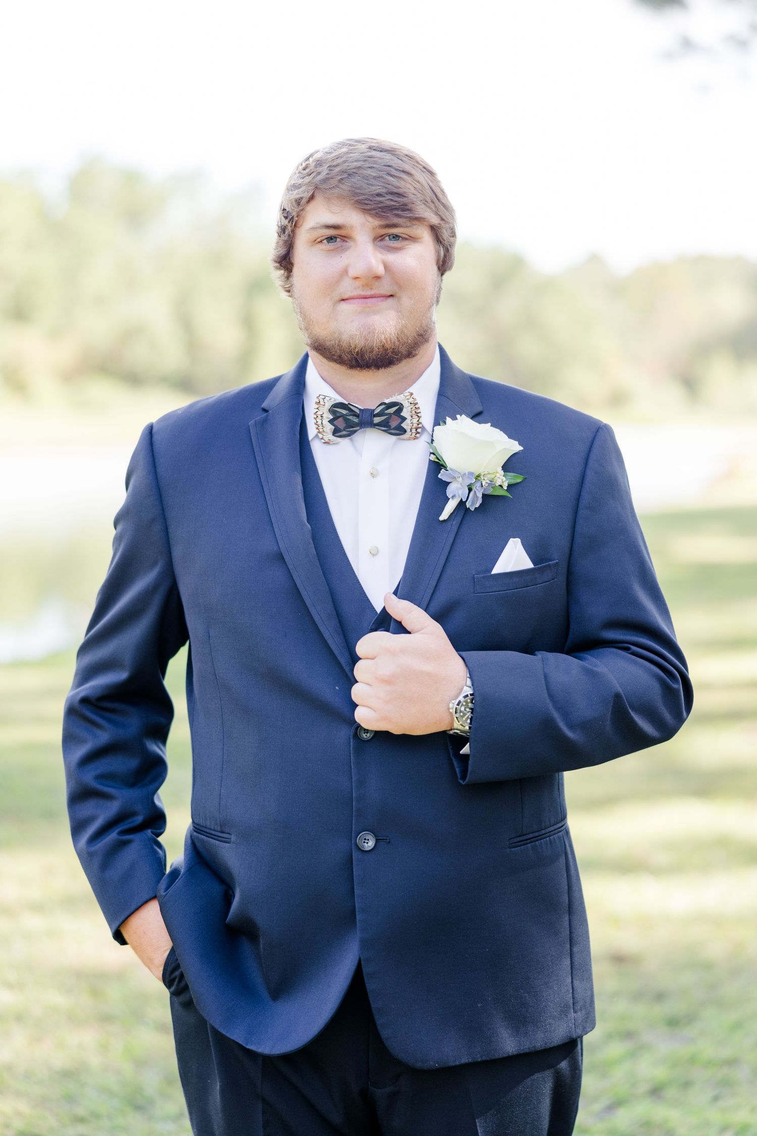 Groom in navy suit with white boutonniere and bowtie poses outdoors on wedding day.