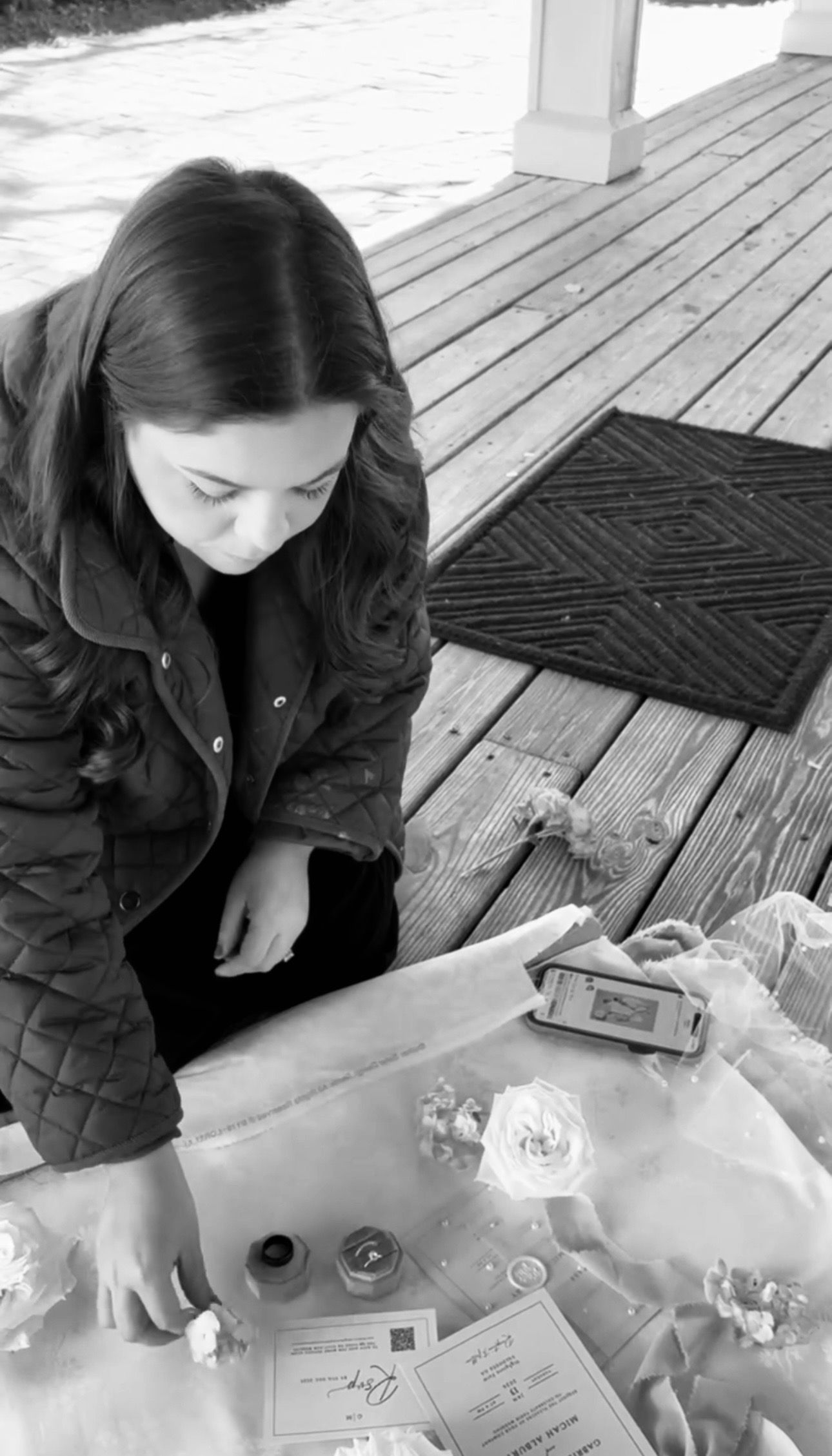 Black and white photo of a woman in a quilted jacket examining items on a table on a wooden deck.