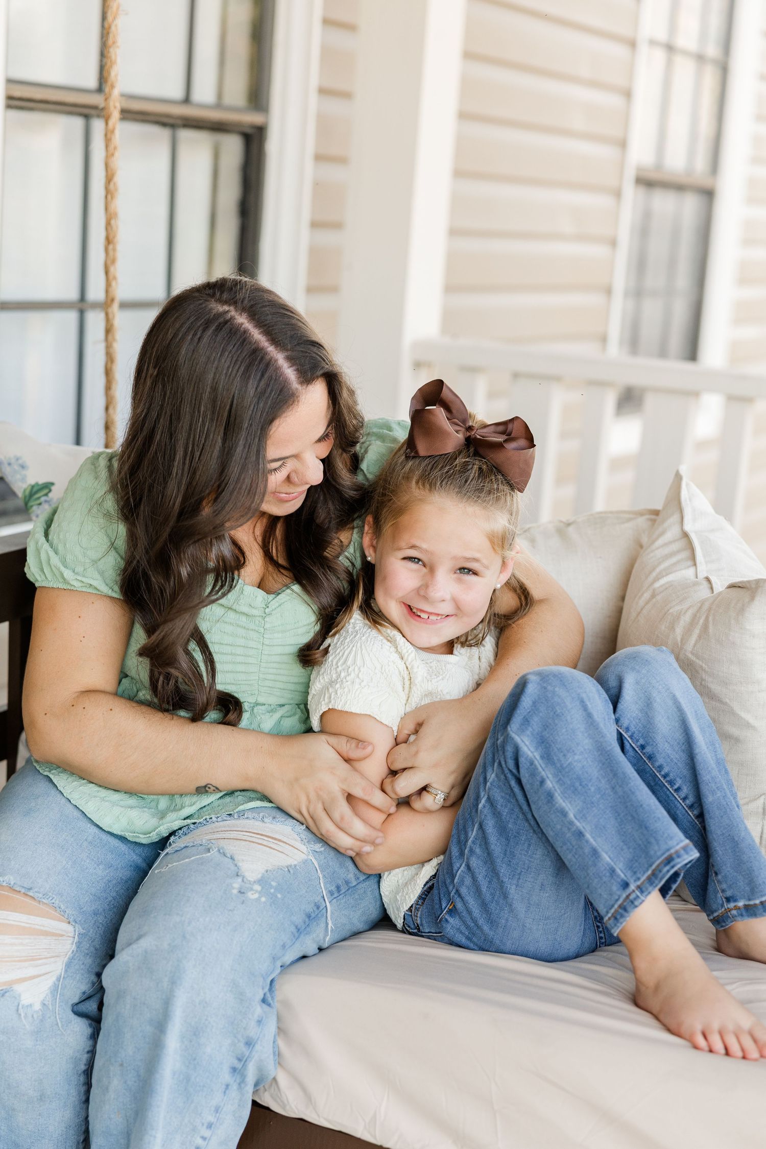 A mother and young daughter share a joyful moment on a porch swing, both wearing jeans and smiling warmly.