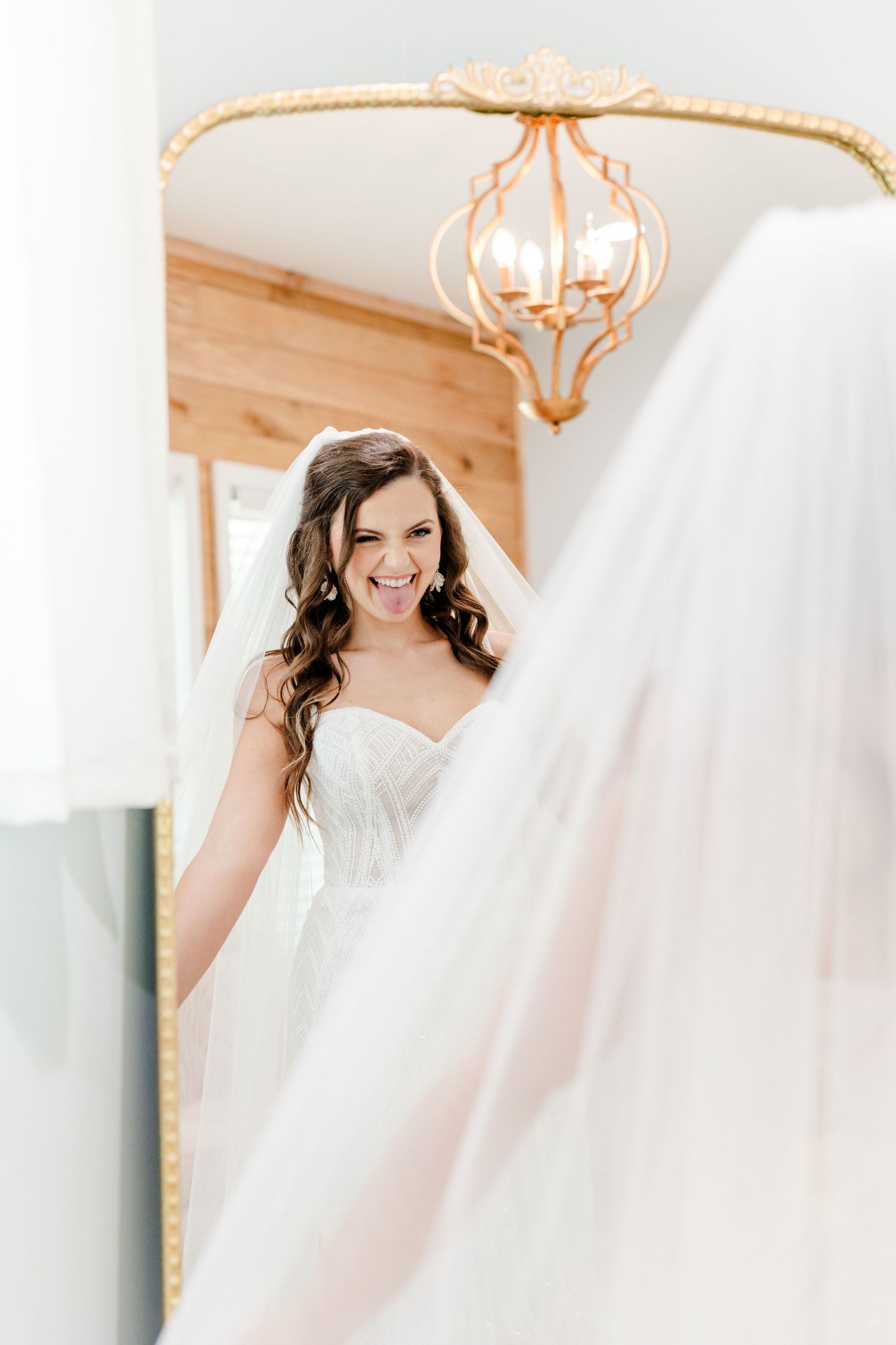 Smiling bride in a strapless white gown and veil stands before a gold-framed mirror in a bright bridal suite.