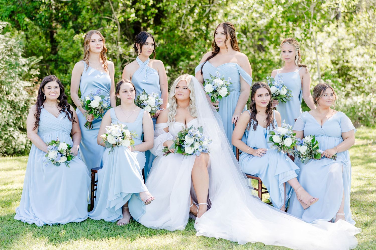 Bridesmaids in light blue dresses pose together on grass with floral bouquets.