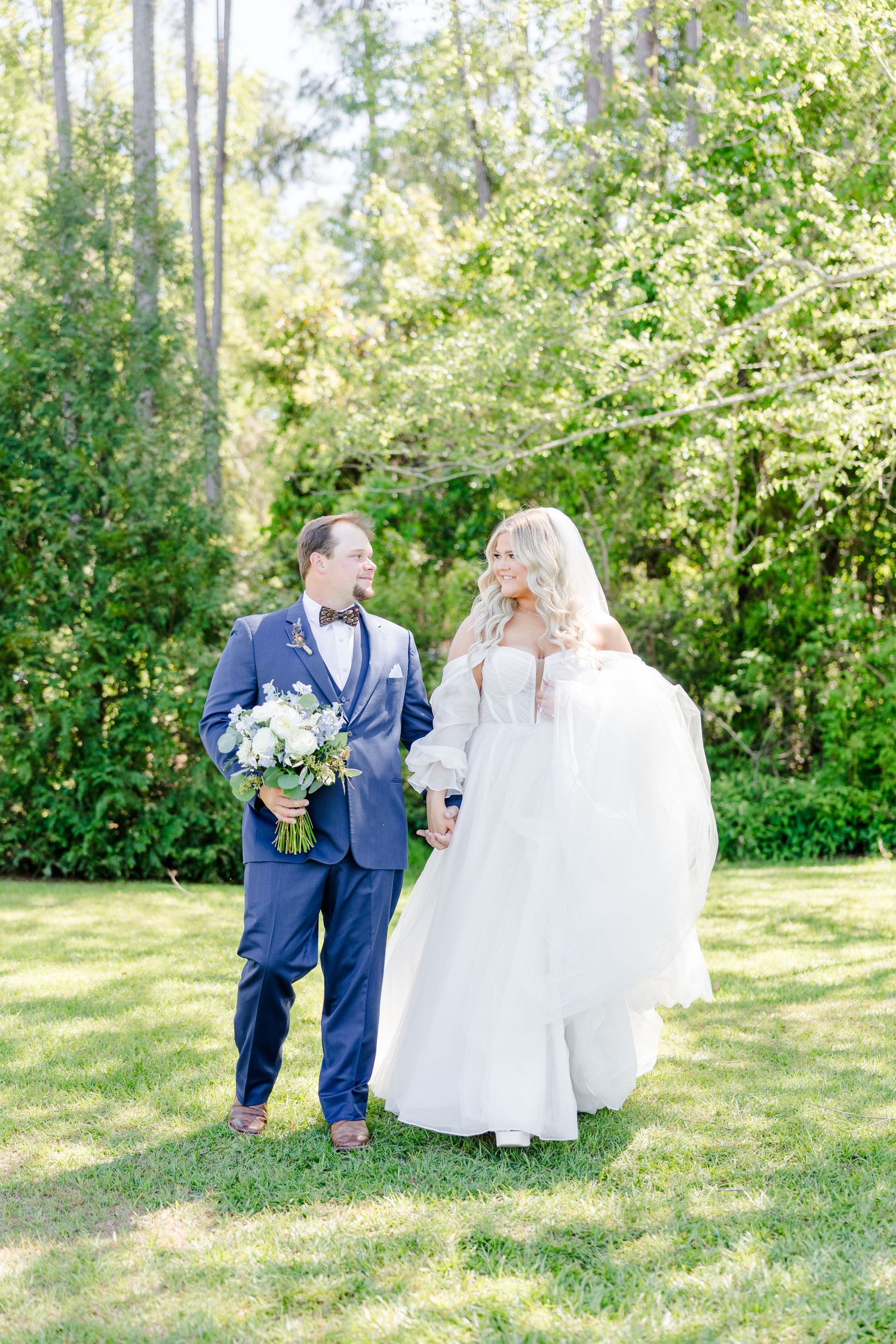 Wedding couple walks together on grassy lawn surrounded by lush spring greenery.