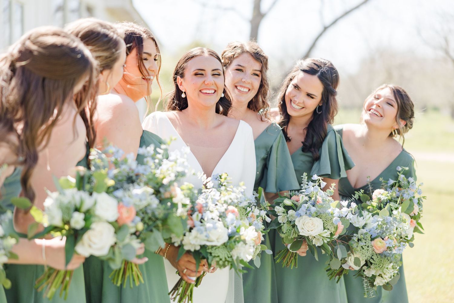 Bridesmaids in sage green dresses hold white floral bouquets at an outdoor wedding.