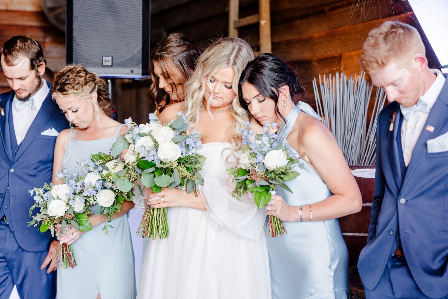 Wedding celebration with bridal party in light blue dresses and navy suits dancing in a rustic barn setting.