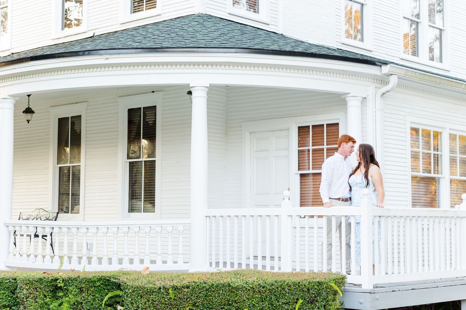 Couple embracing on white wraparound porch of elegant Victorian home during outdoor engagement photo session.