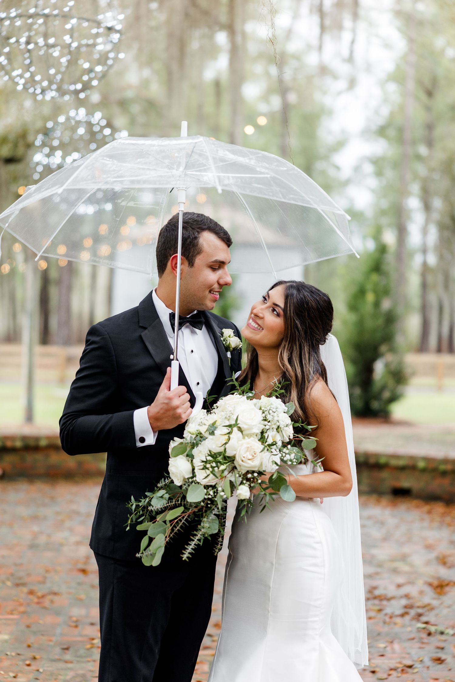 Bride and groom share a romantic moment under a clear umbrella in a rainy outdoor wedding setting with white bouquet.