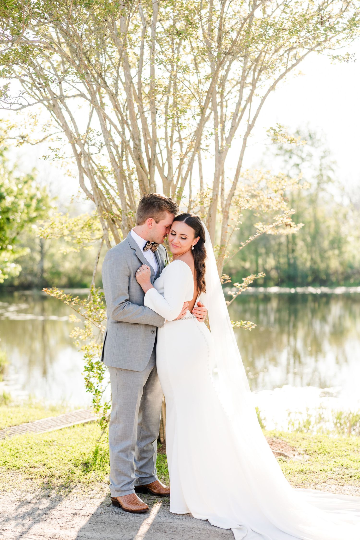 Wedding couple embraces by lakeside with sunlit trees in background.