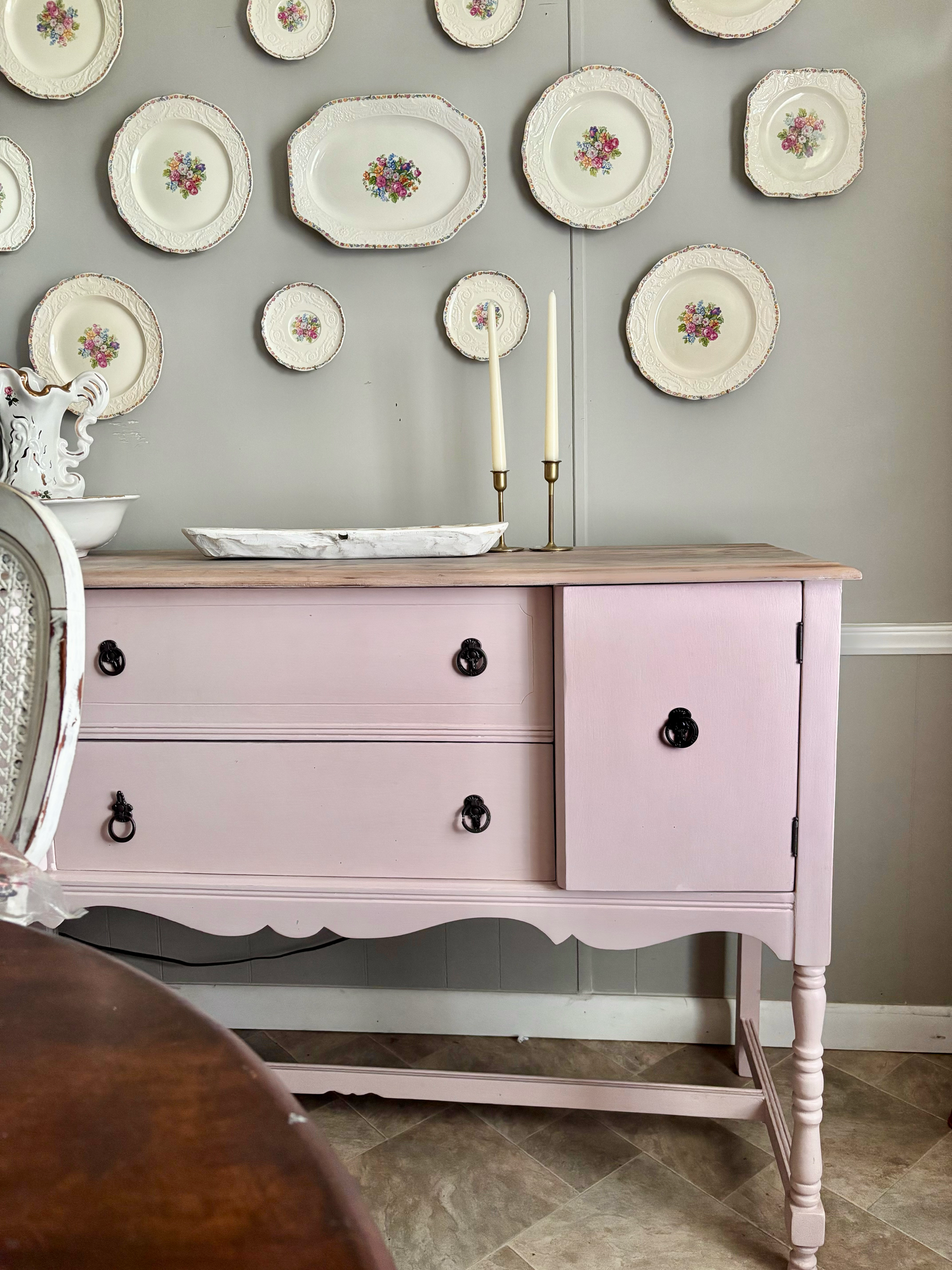 Pink painted vintage buffet sideboard with black hardware, wood top, displayed with decorative plates on gray wall.