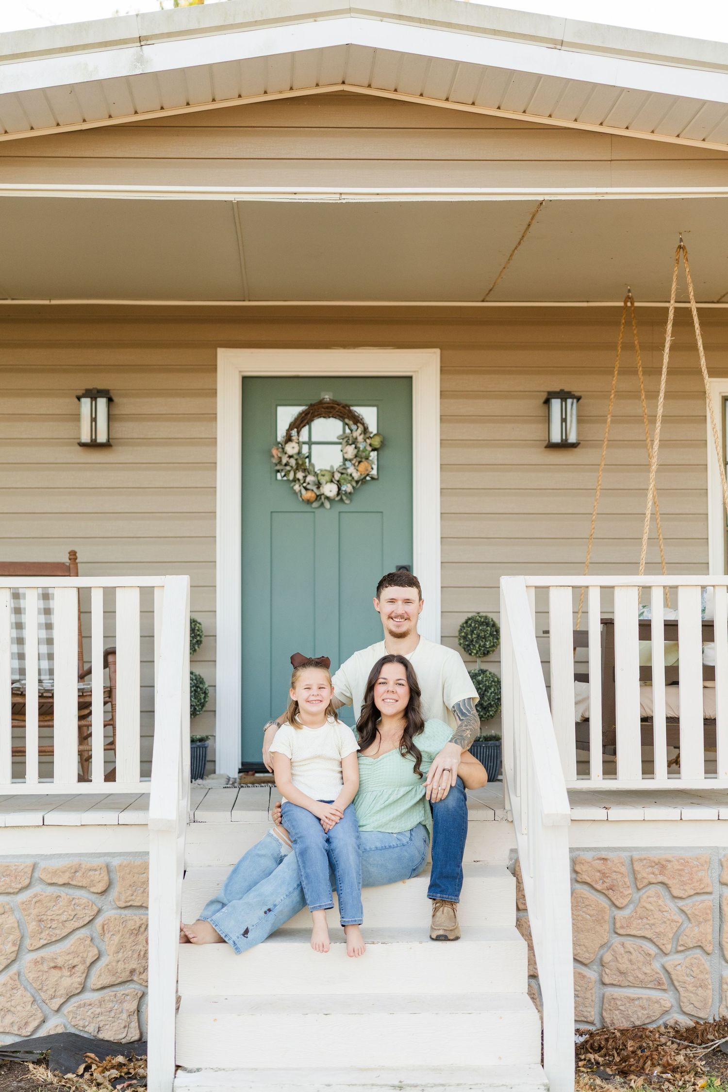 Happy family of three sitting on front porch steps of a cozy home with teal door and wreath.
