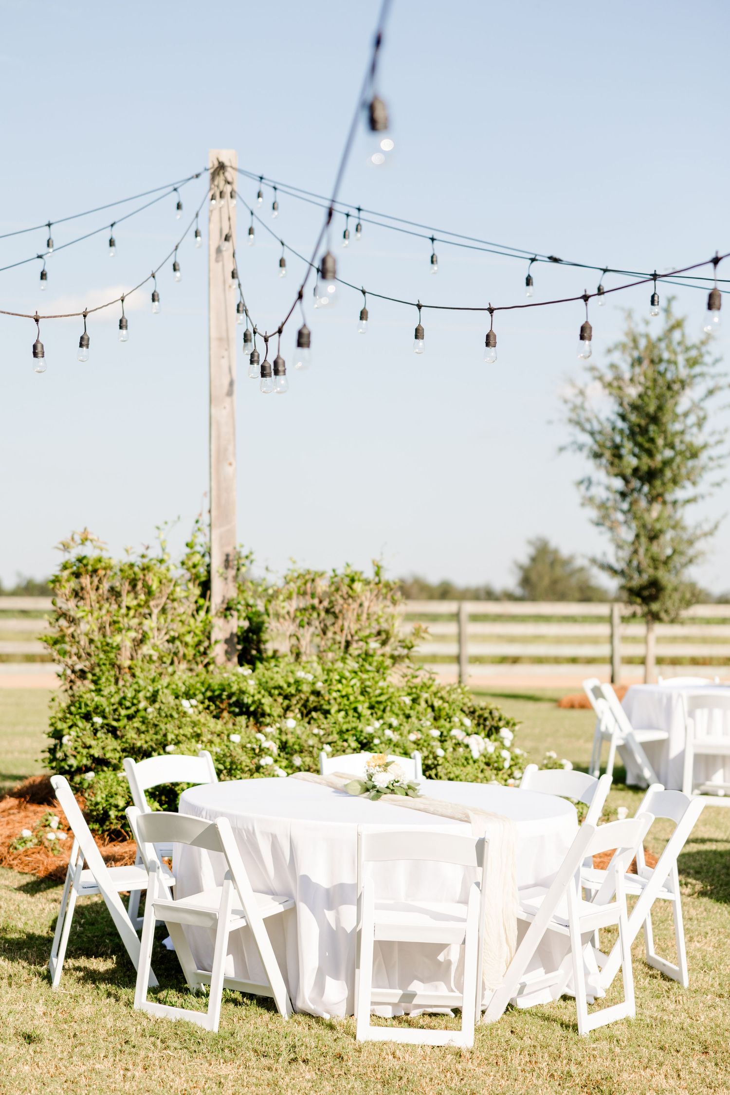 Outdoor wedding reception setup with white chairs and string lights in a garden setting.