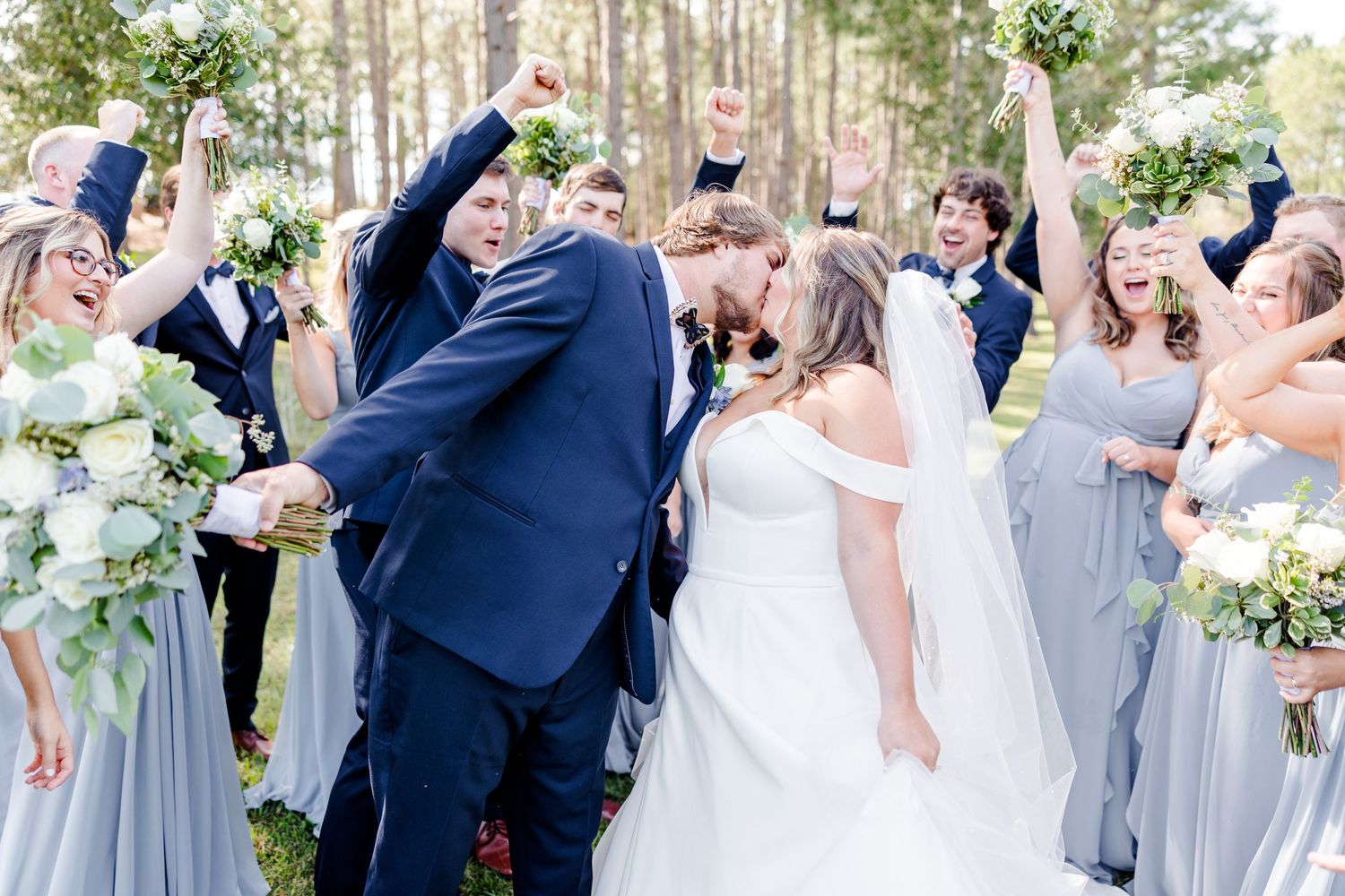 Wedding party celebrates joyfully during outdoor photo session among pine trees.