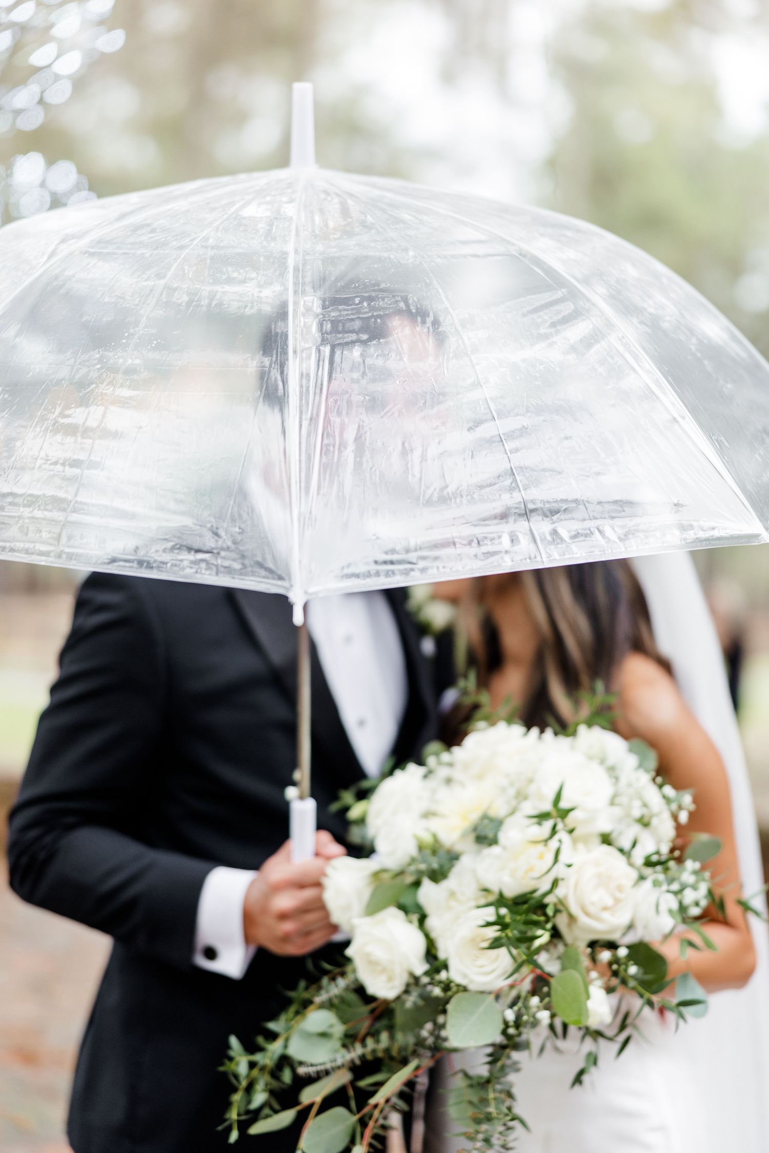 Bride and groom share a romantic moment under a clear bubble umbrella, bride holding a white and greenery bouquet.