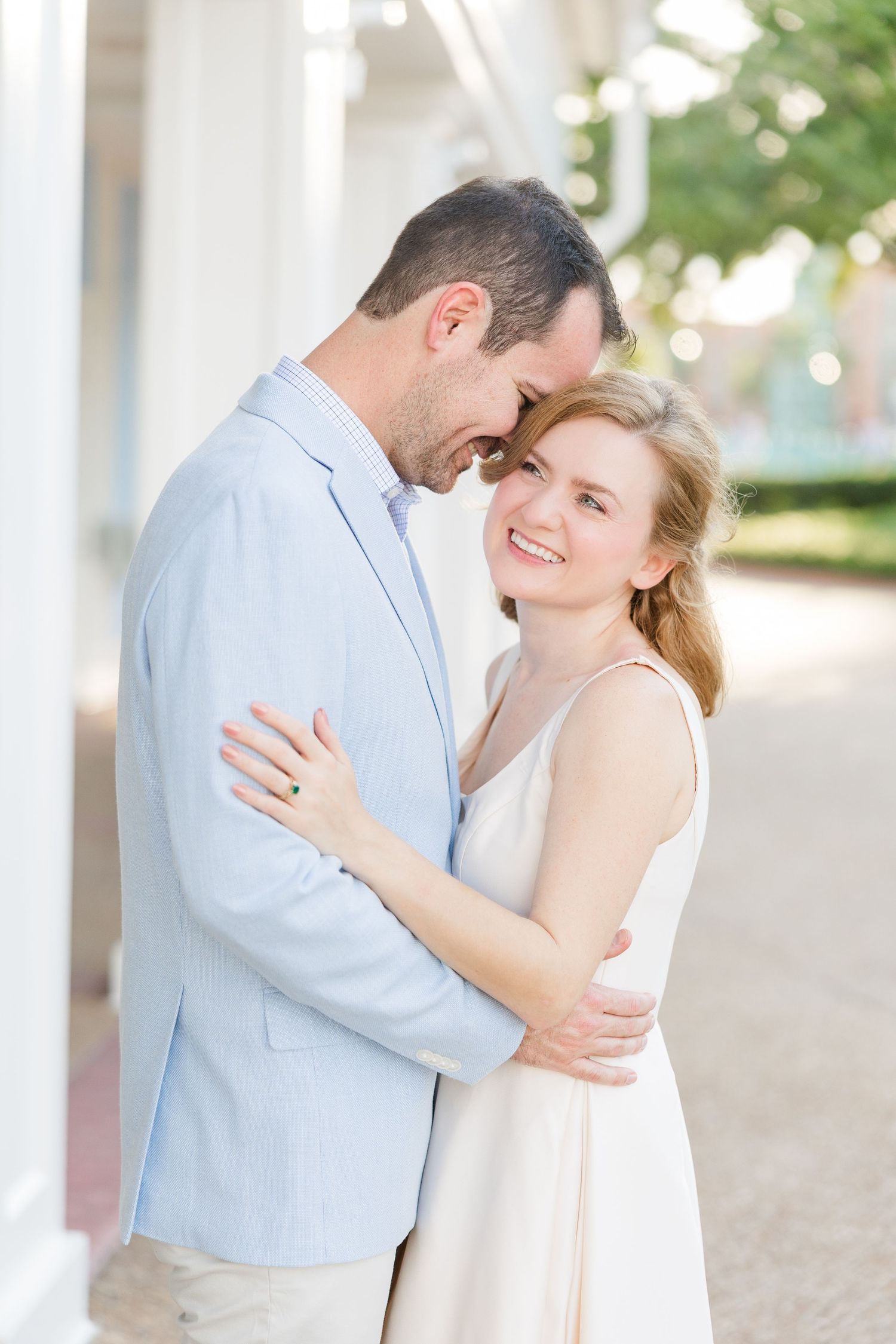 A smiling couple embraces outdoors, woman in white dress, man in light blue blazer, sharing a tender romantic moment.