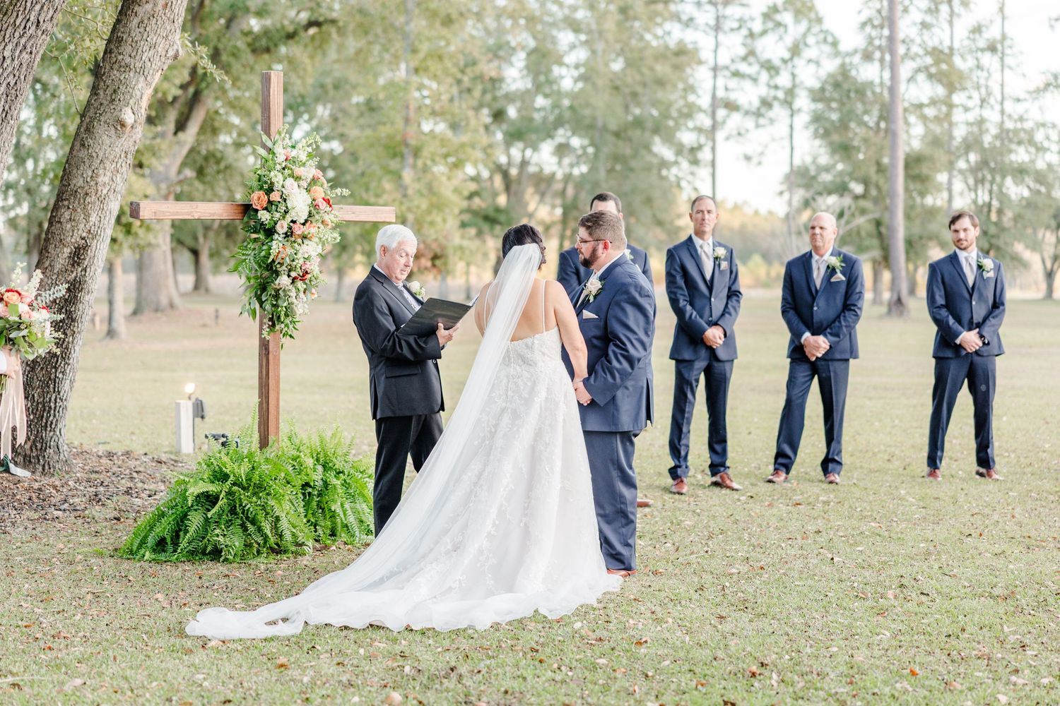 Wedding ceremony setup with guests and wedding party gathered under towering trees.