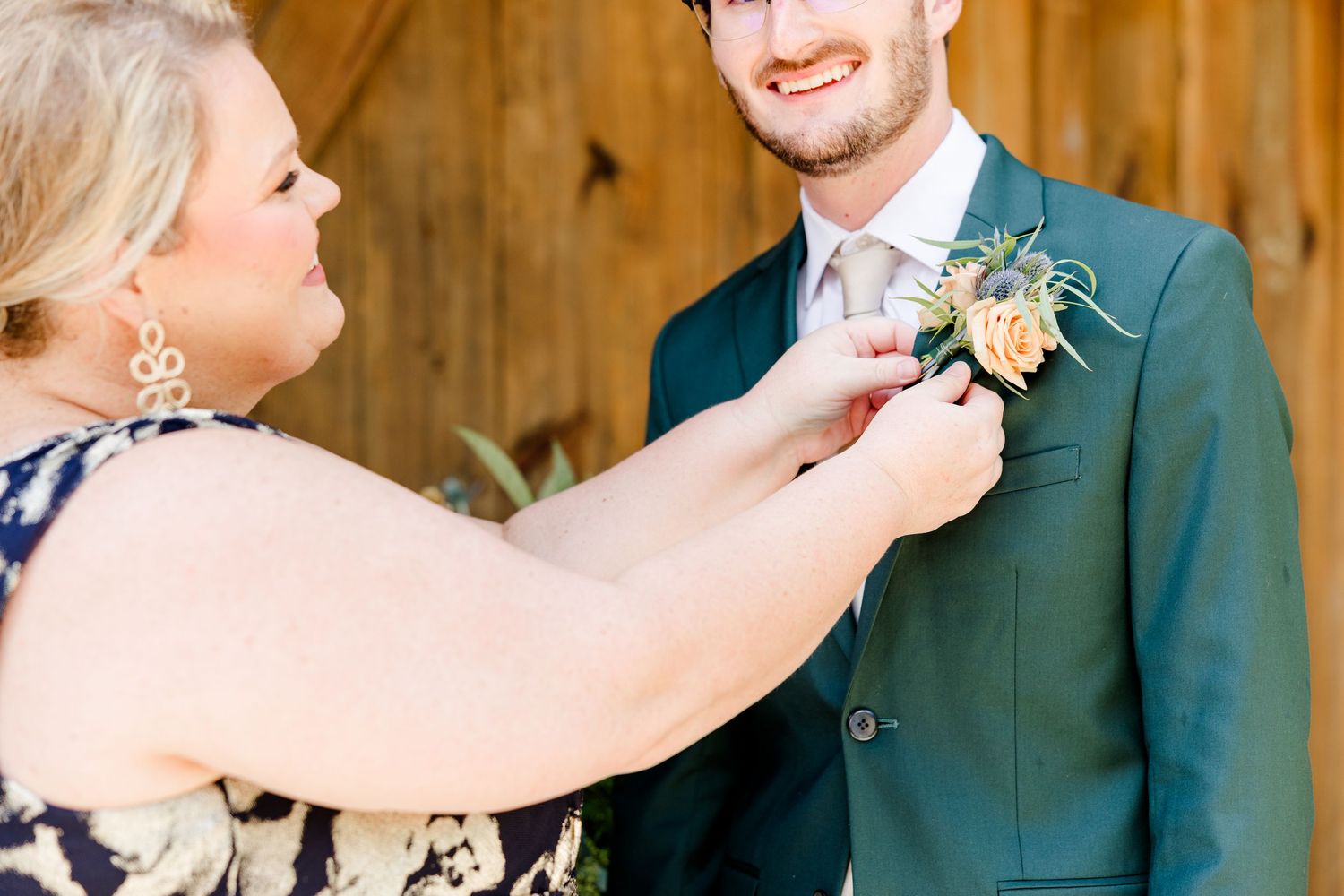 Woman pinning a floral boutonniere on a smiling groom in a dark suit at a rustic barn wedding.