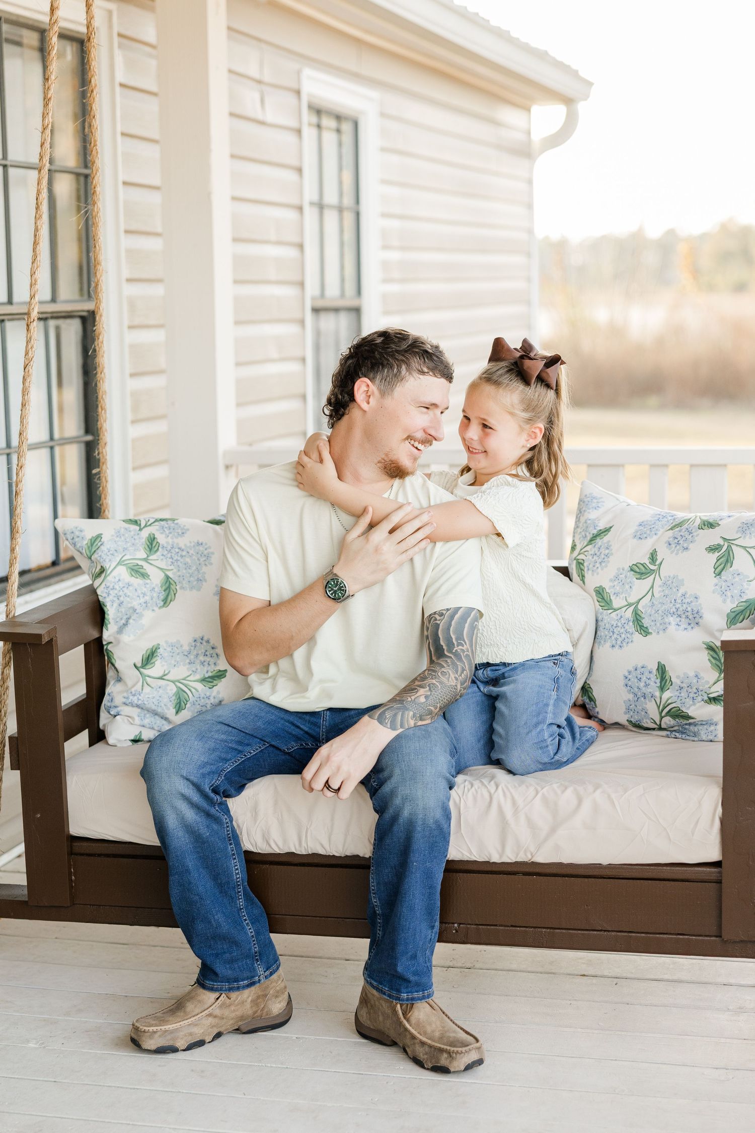 A father and daughter share a warm embrace on a porch swing, both wearing jeans and white shirts.