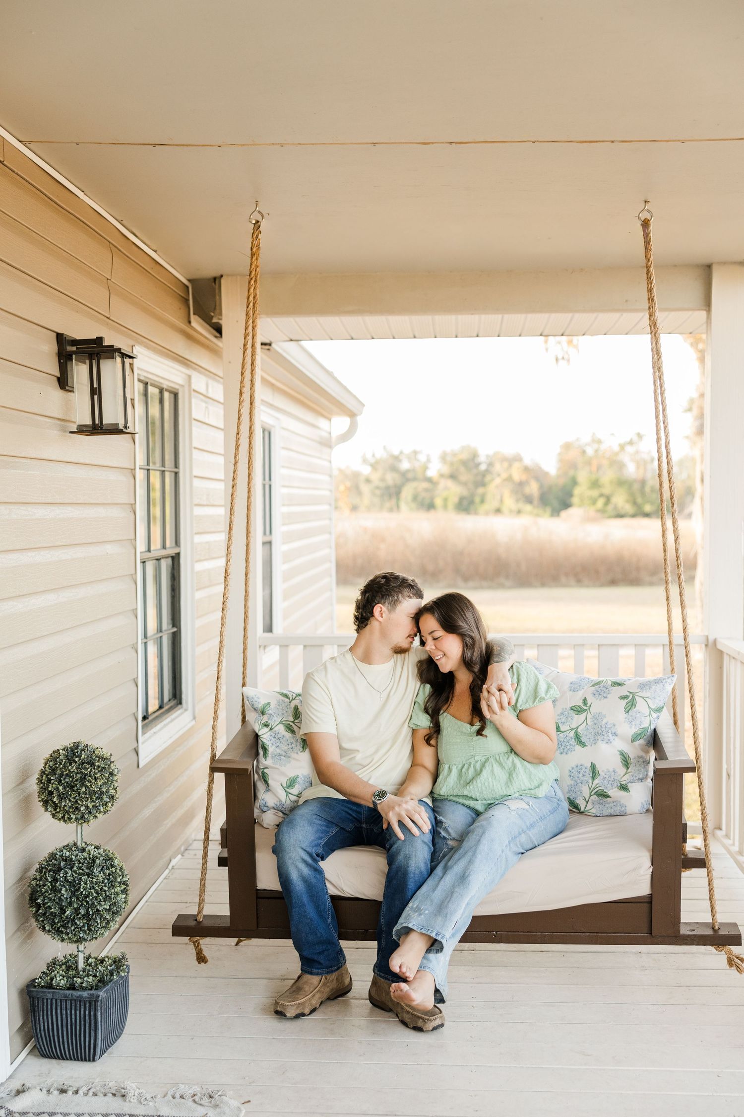 Couple sitting close on a porch swing with floral pillows, surrounded by golden fields at sunset.