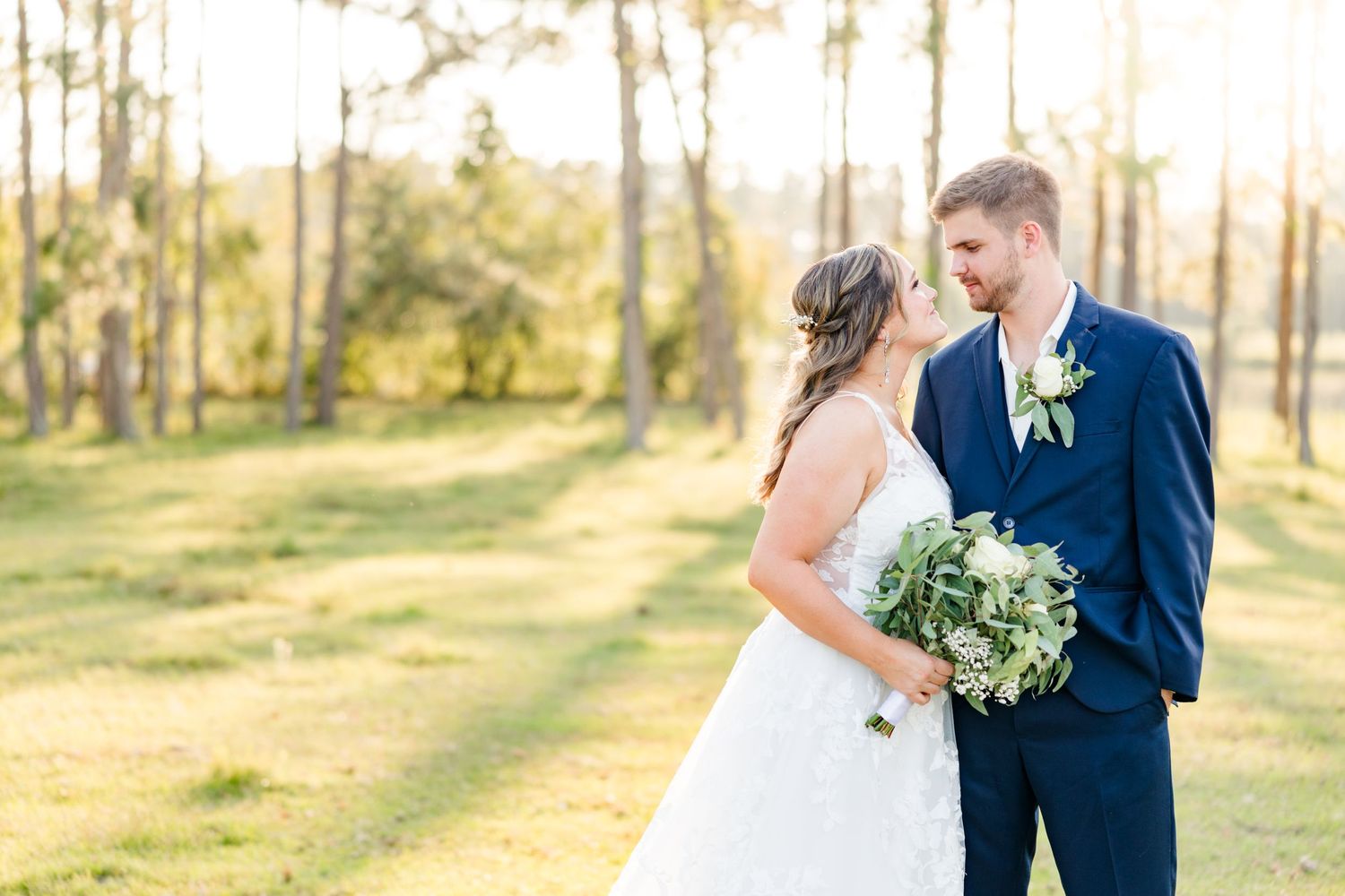 A wedding couple poses together in a pine forest during sunset holding a white and green bouquet.