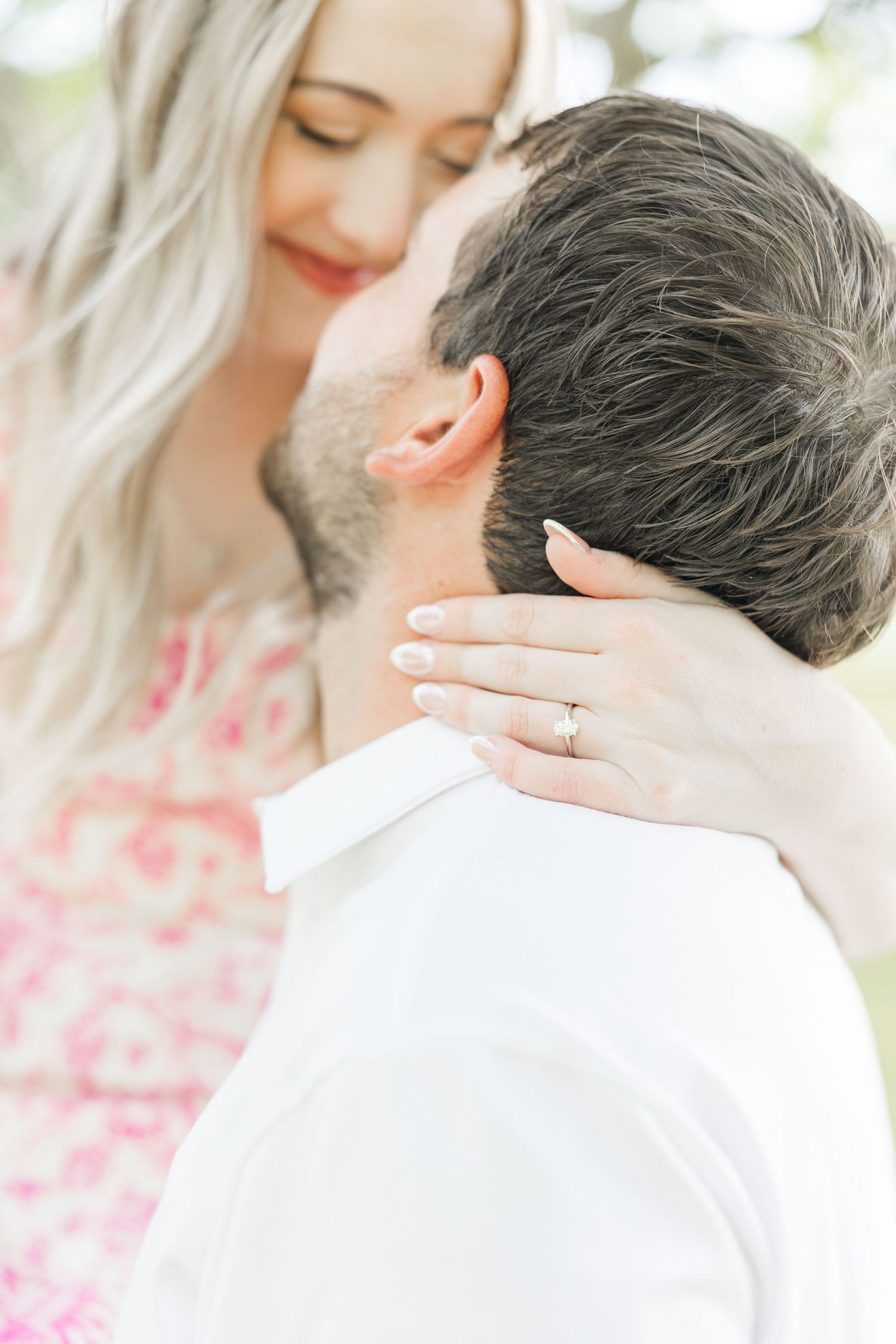Blonde woman in floral dress embraces man in white shirt, showcasing engagement ring while sharing tender moment outdoors.