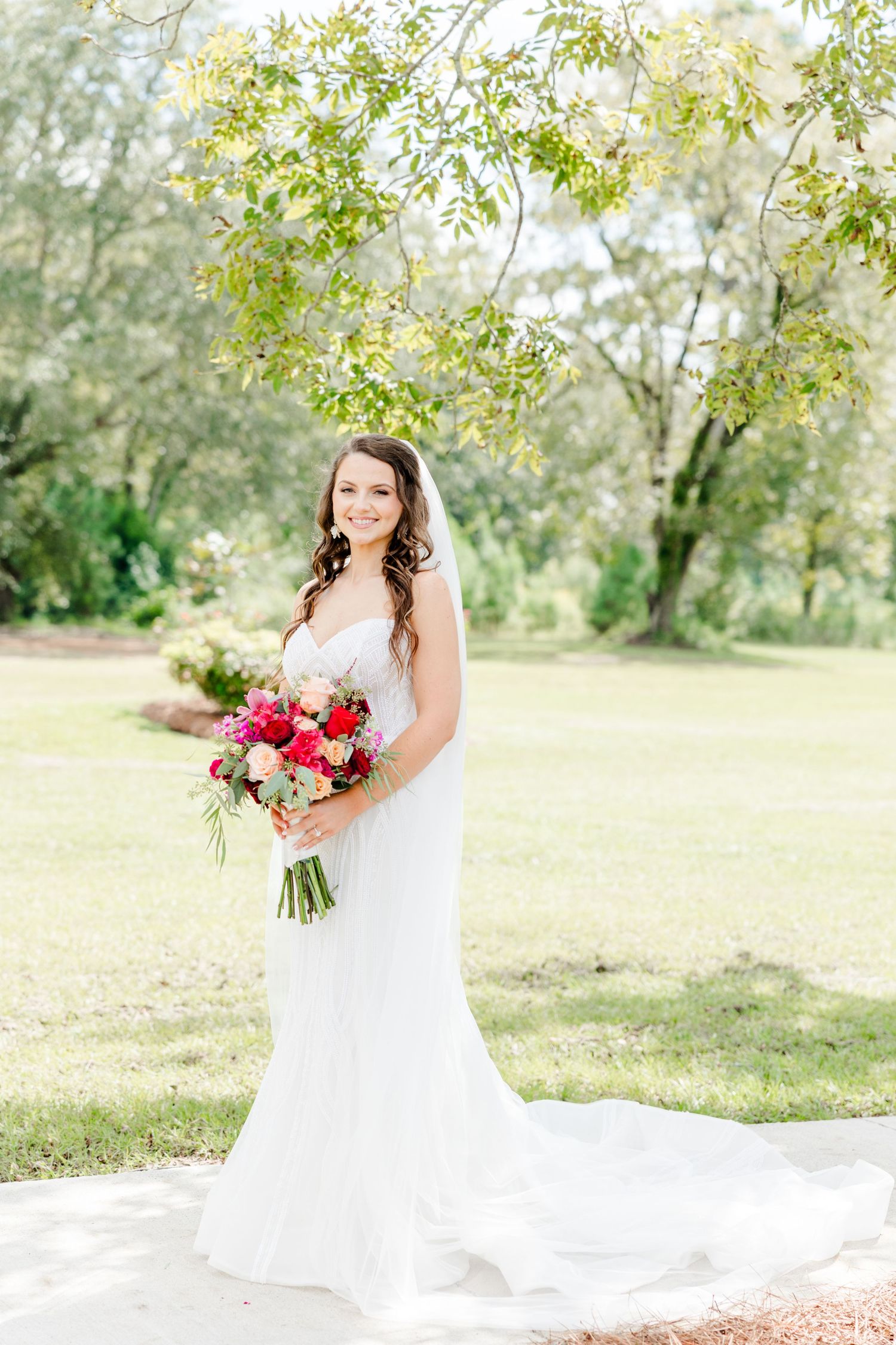 Bride in white gown holding vibrant pink bouquet stands under green trees on sunny day.
