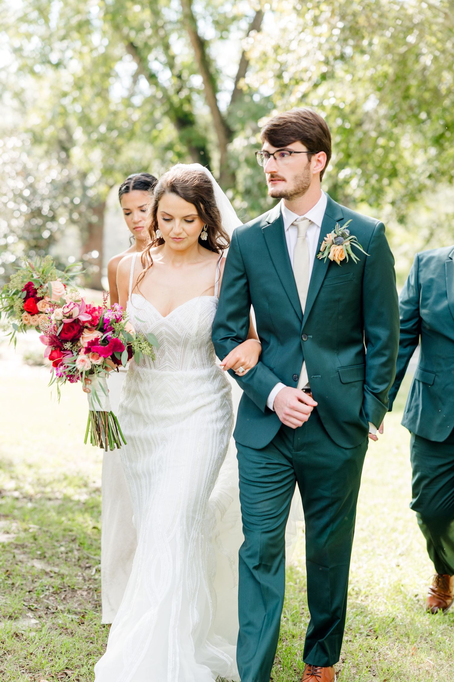 Bride in white gown holding red bouquet walks arm-in-arm with groom in dark green suit outdoors.