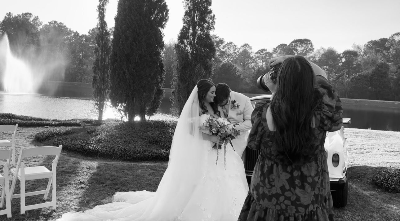 Black and white wedding photo of bride being photographed near fountain and trees at outdoor ceremony.