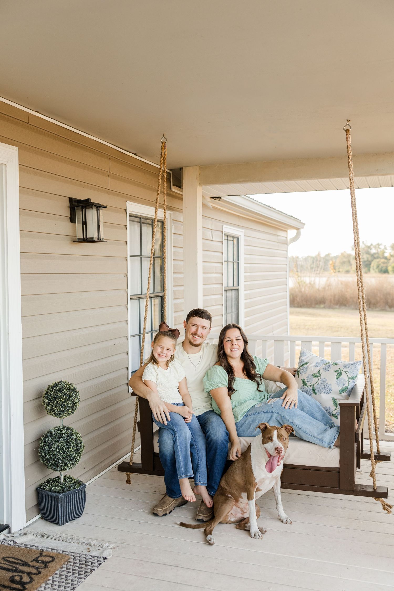 Family of three with dog relaxing on porch swing at farmhouse home, smiling couple with young daughter outdoors.