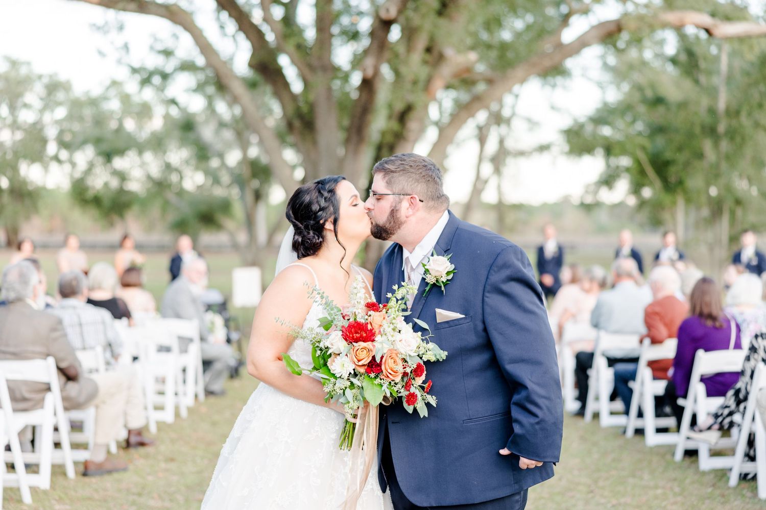 Wedding ceremony site with white chairs and floral arrangements lined up under trees.