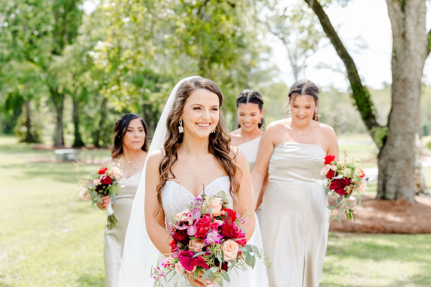 Bridesmaids in elegant white dresses holding vibrant pink and red bouquets outdoors.