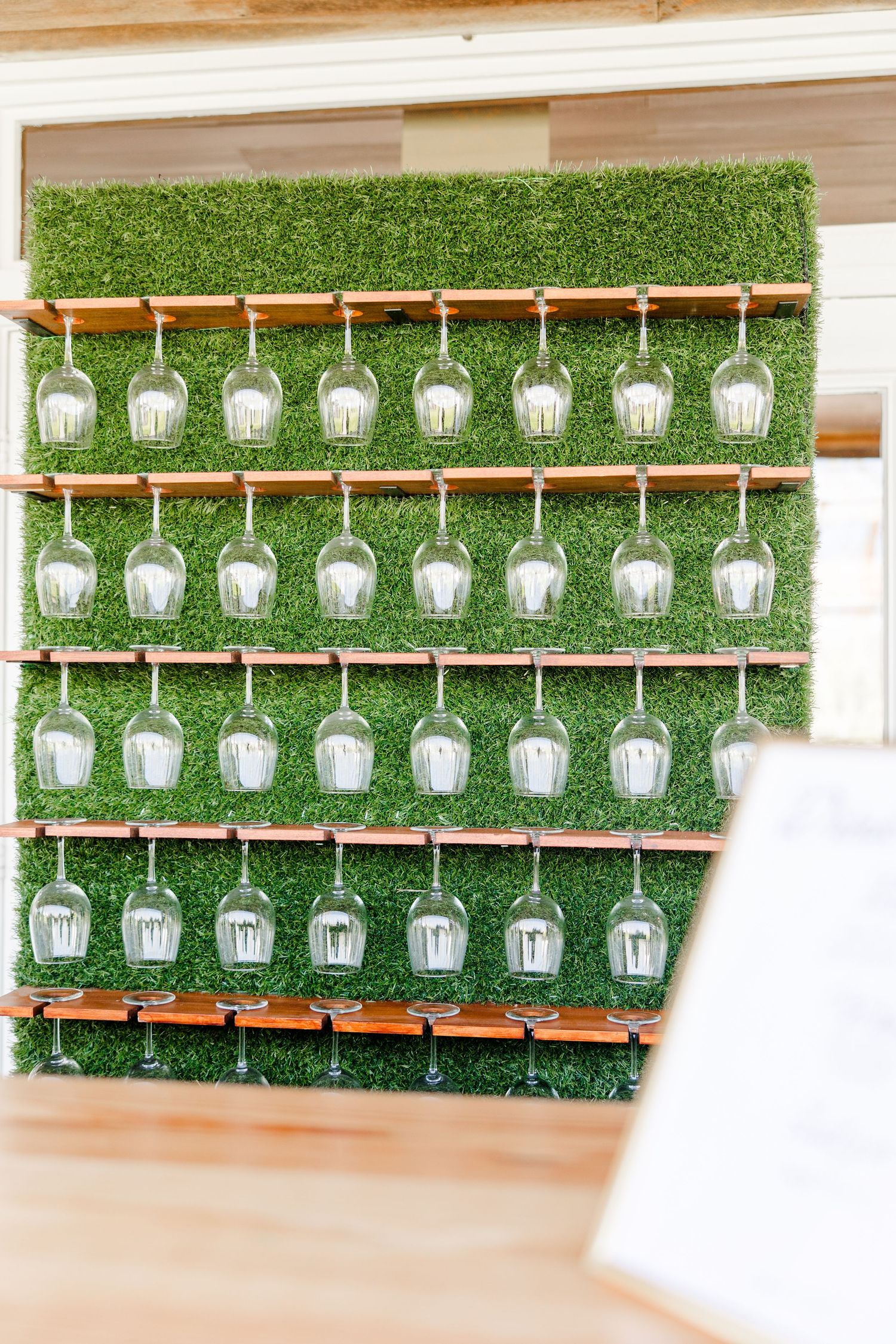 A creative wedding display featuring rows of wine glasses arranged on a green hedge wall.