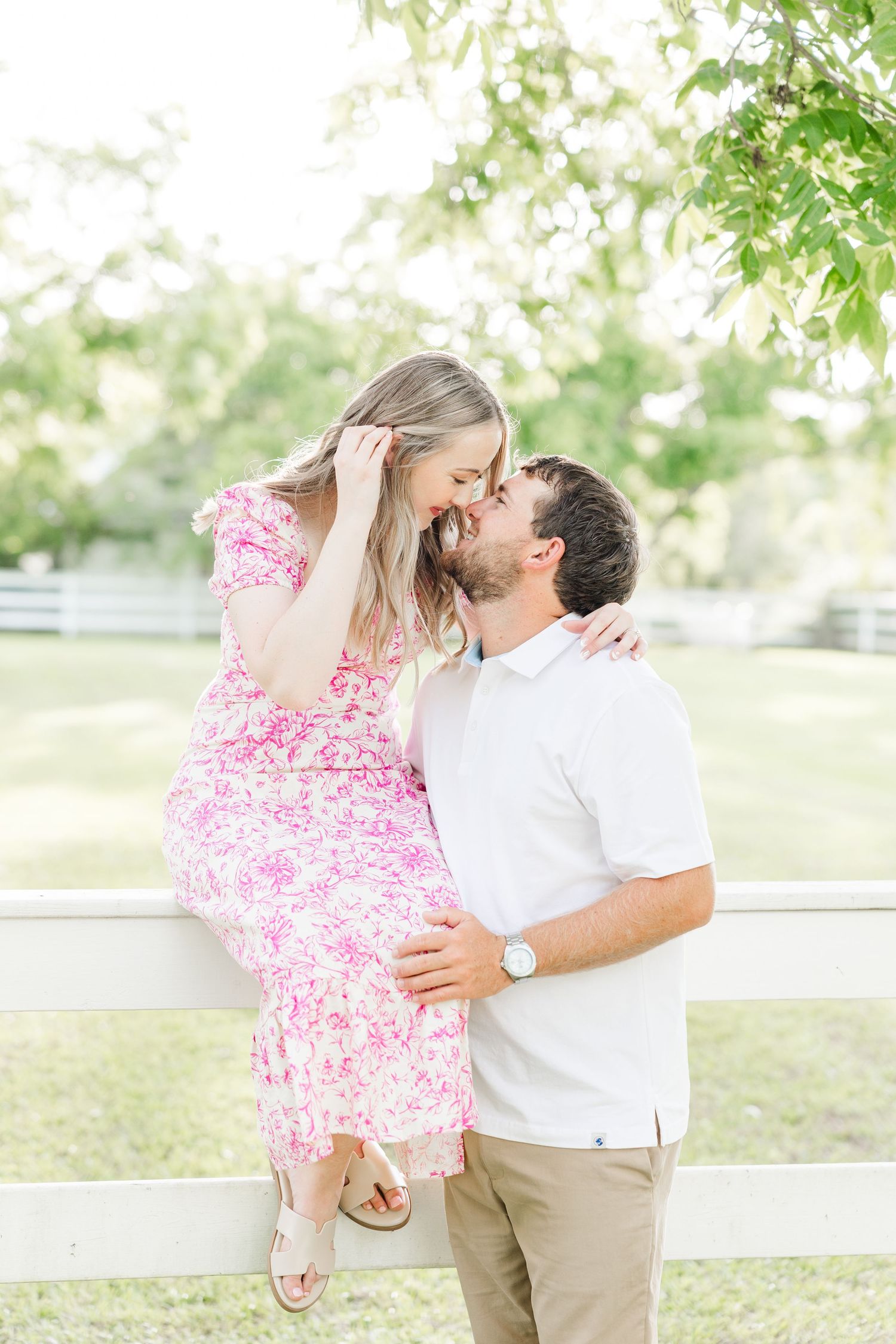 A couple shares a romantic kiss outdoors, woman in pink floral dress, man in white polo, leaning against a white fence.