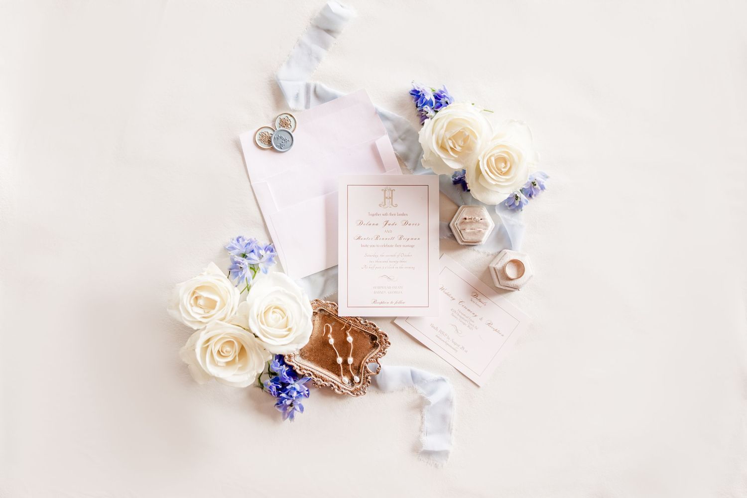 Wedding flat lay with cream roses, blue flowers, and invitation cards on white background.