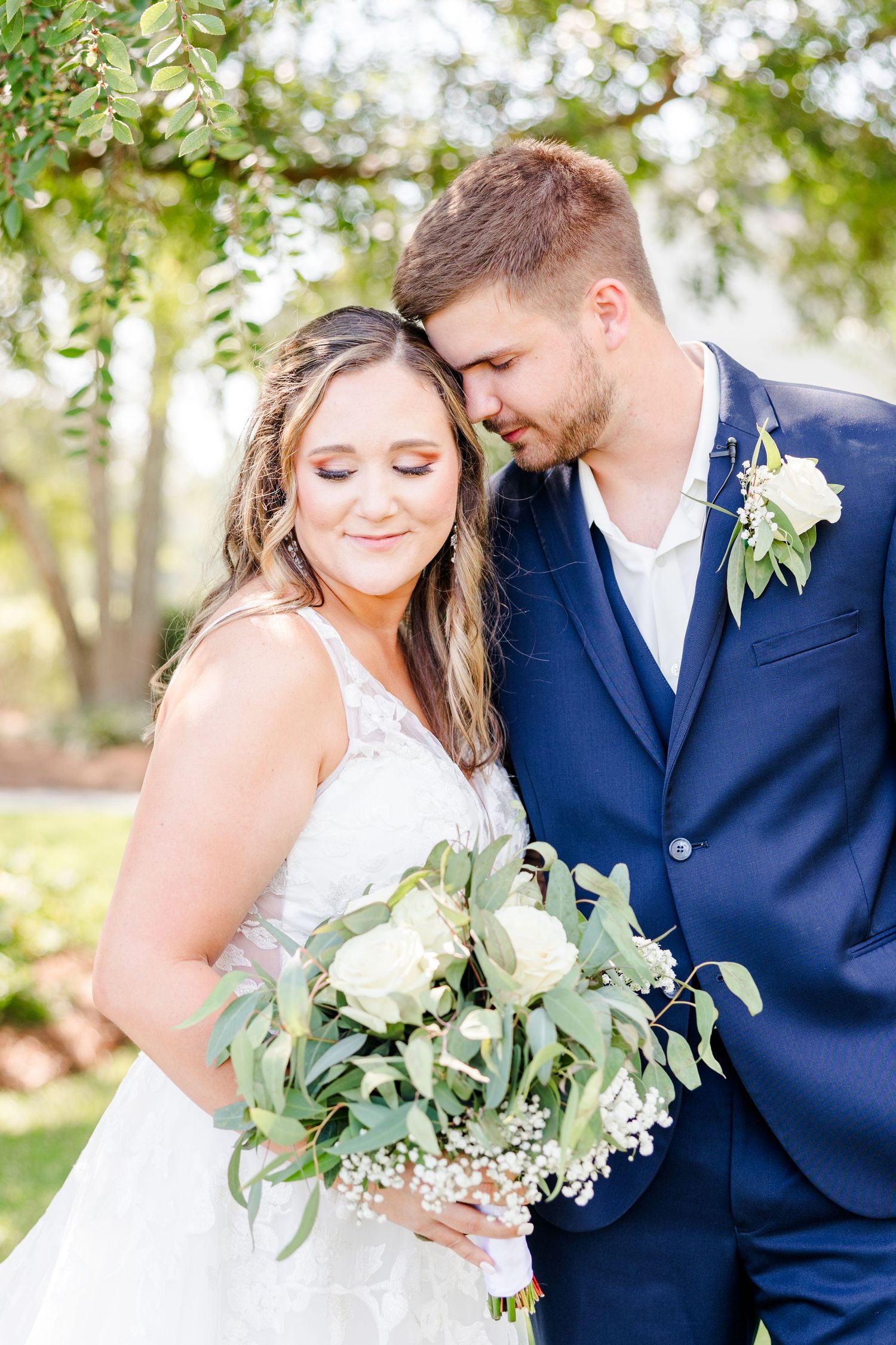 Wedding couple embraces in sunlit garden setting with white floral bouquet and navy attire.