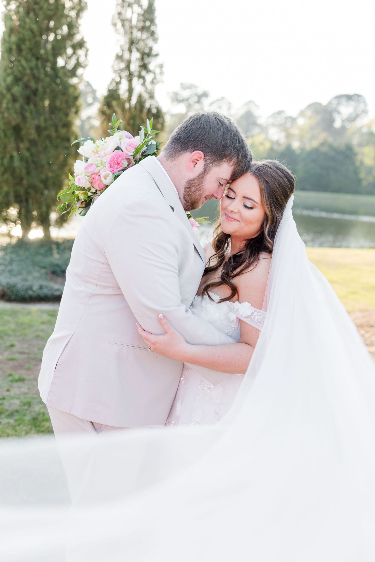 Bride and groom share romantic embrace outdoors, her veil flowing, holding pink bouquet in soft golden light.