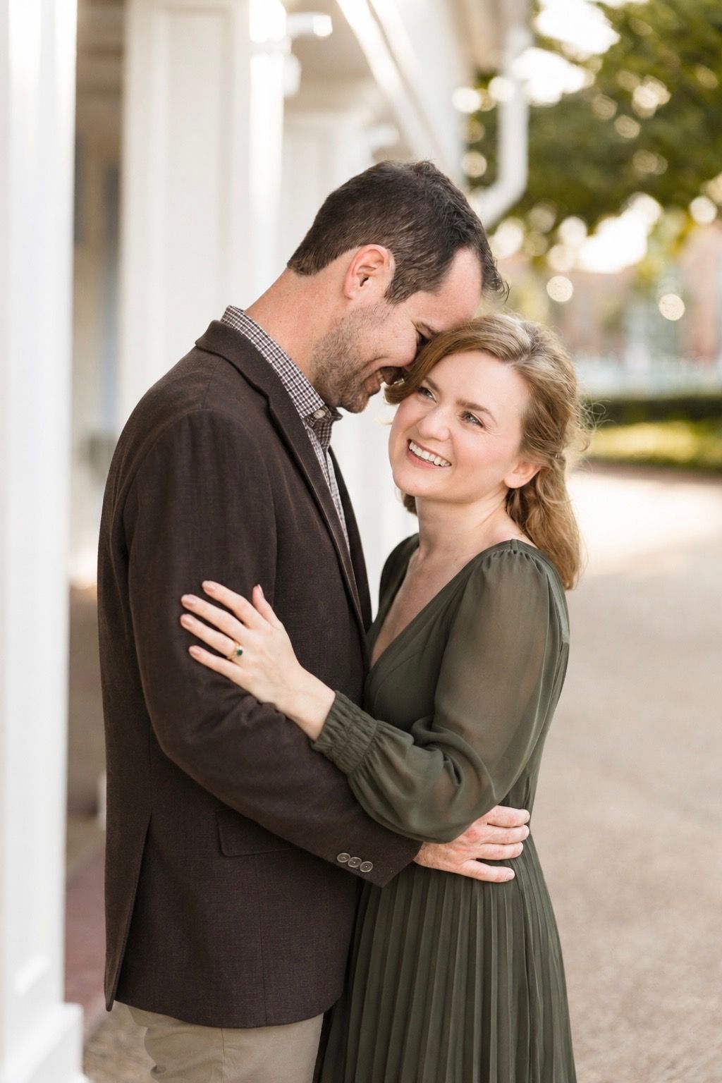 A smiling couple in elegant attire share a romantic forehead-to-forehead embrace outdoors.