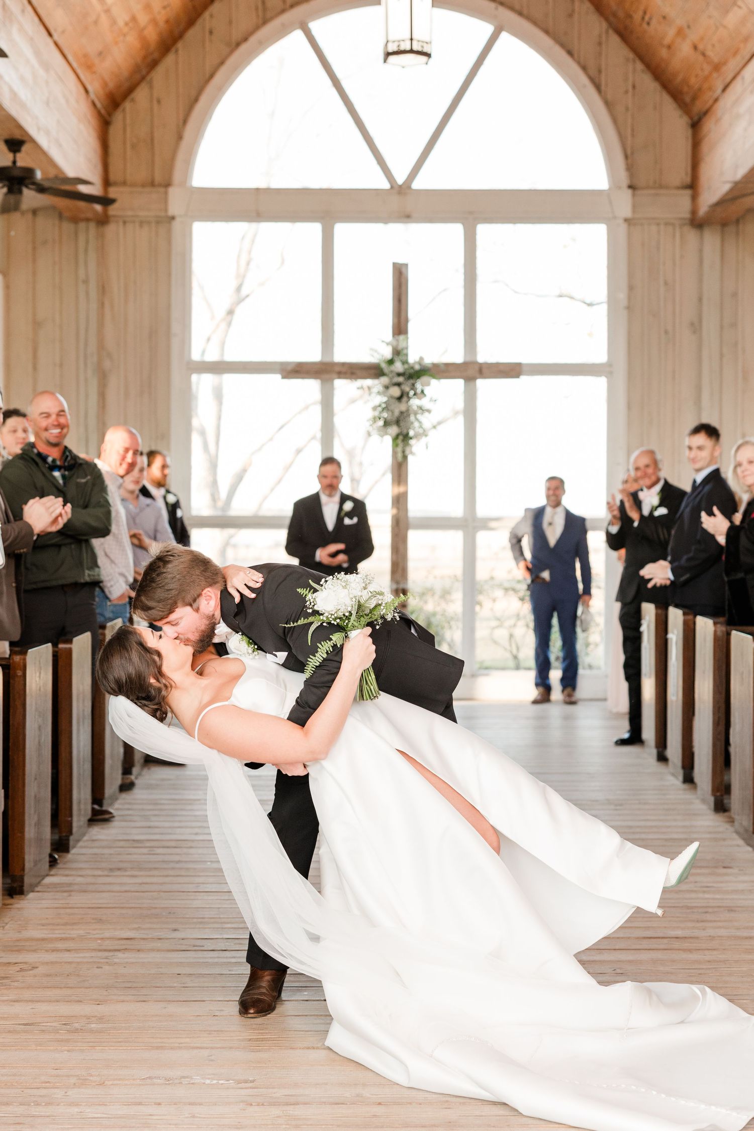 Wedding couple performing dramatic dip pose in wooden chapel with guests watching and applauding.