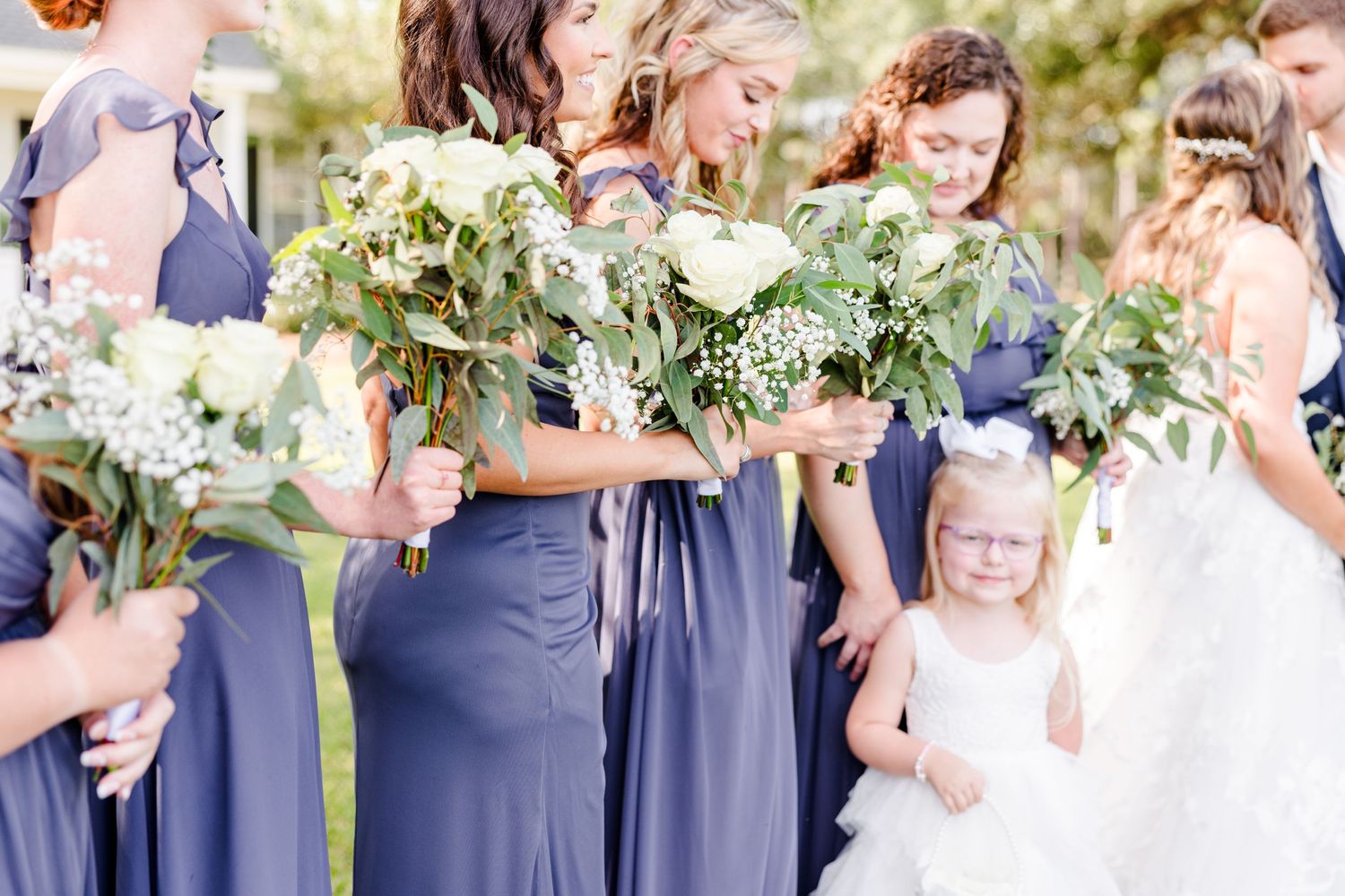Bridesmaids in dusty blue dresses hold white floral bouquets during outdoor ceremony.