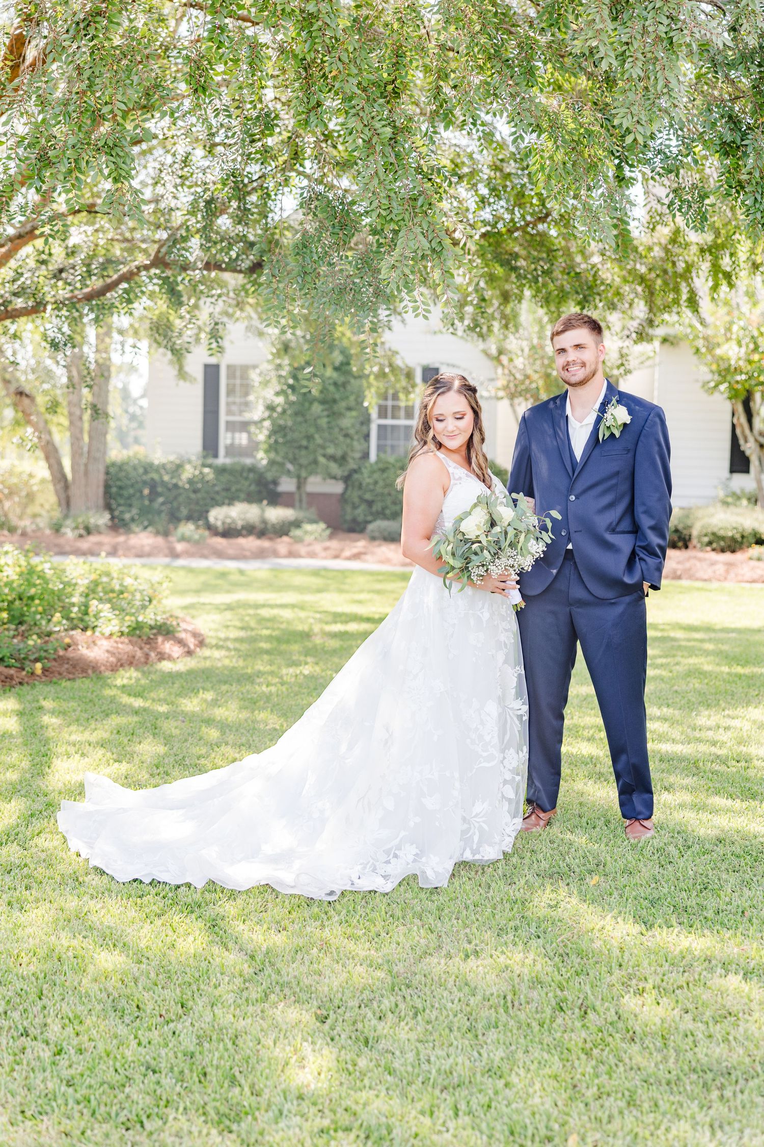 Wedding couple poses beneath leafy trees on manicured lawn with classic house in background.