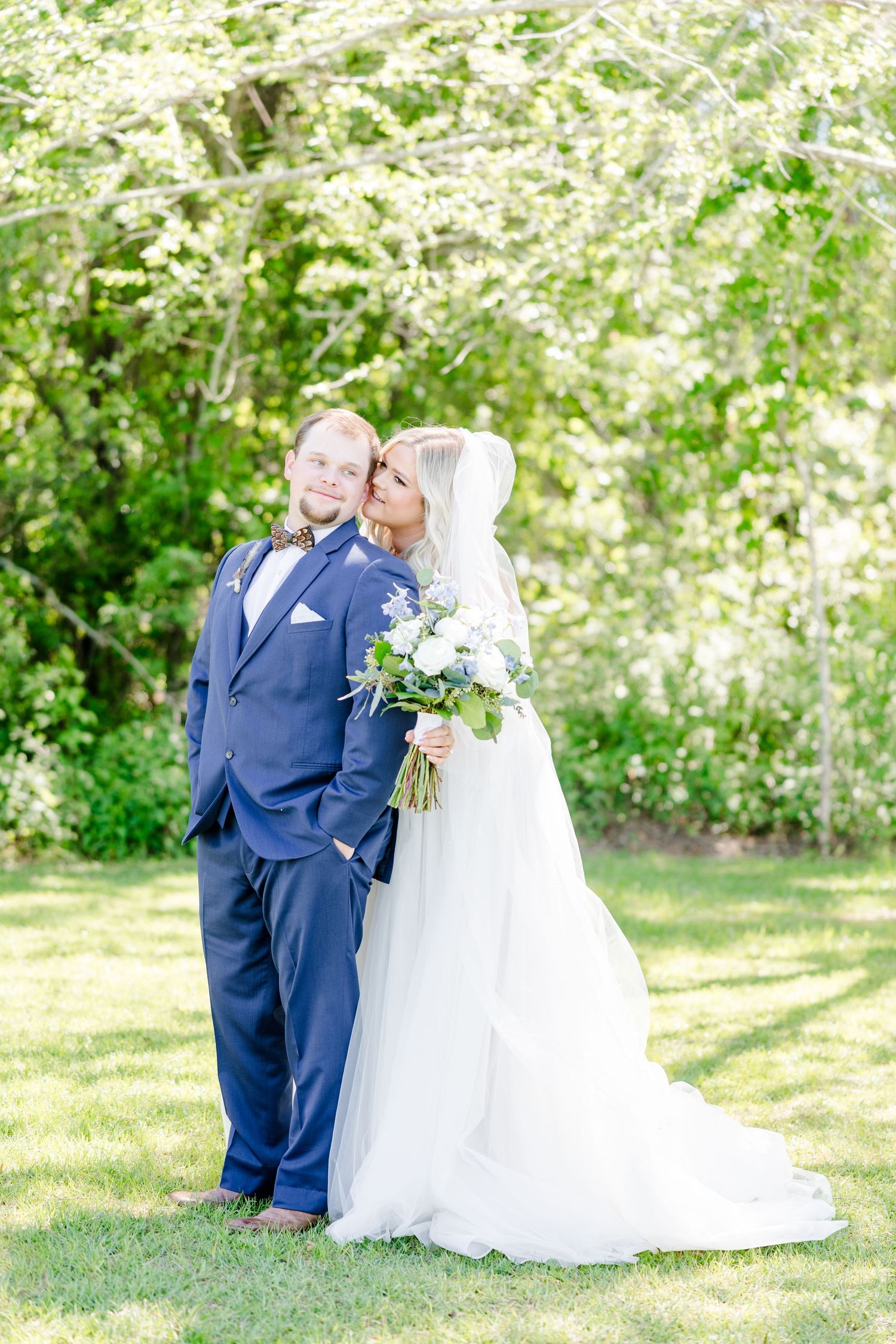 Wedding couple in navy suit and white gown pose together in a sunlit garden setting.