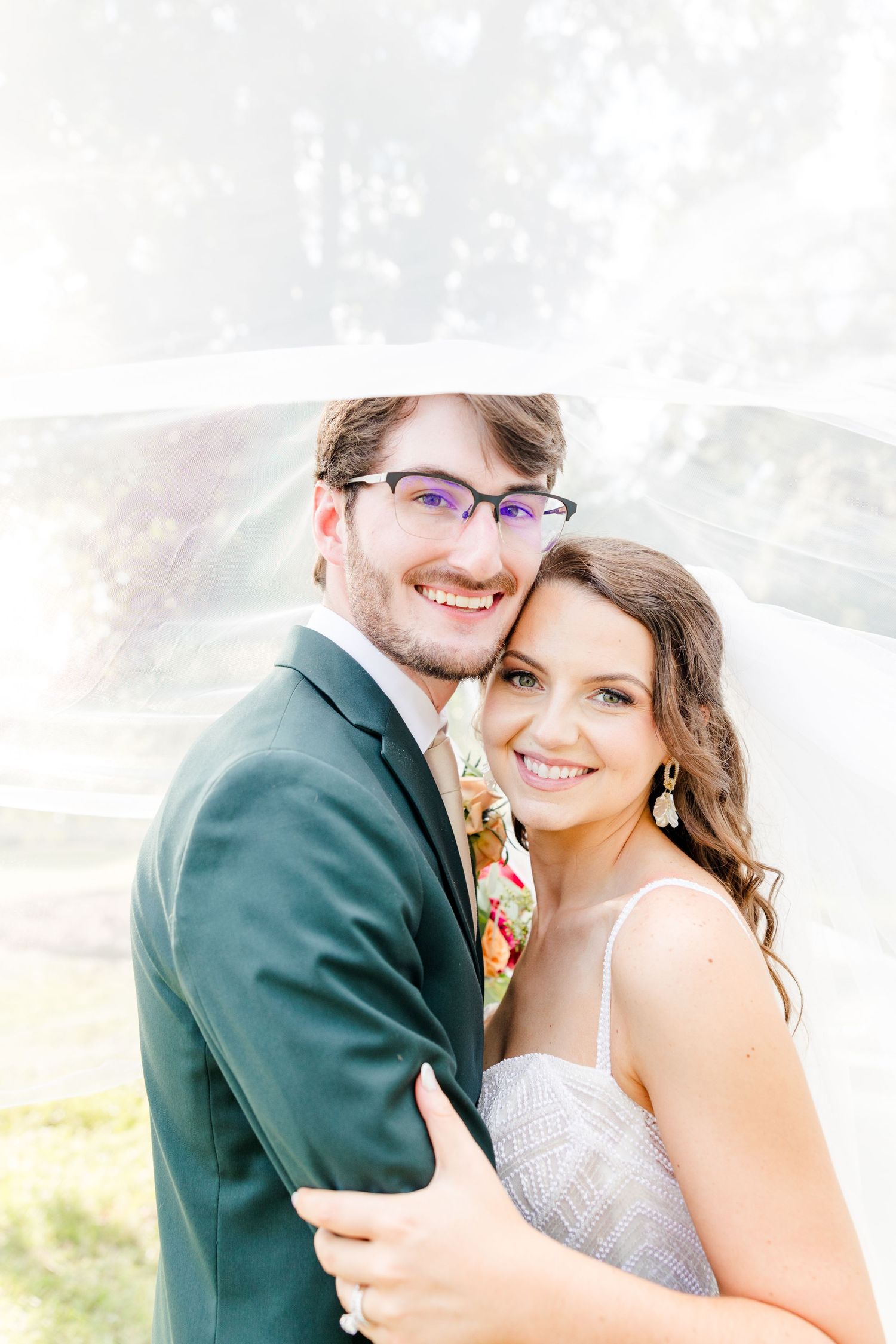 A smiling bride and groom pose under a flowing white veil outdoors on their wedding day.