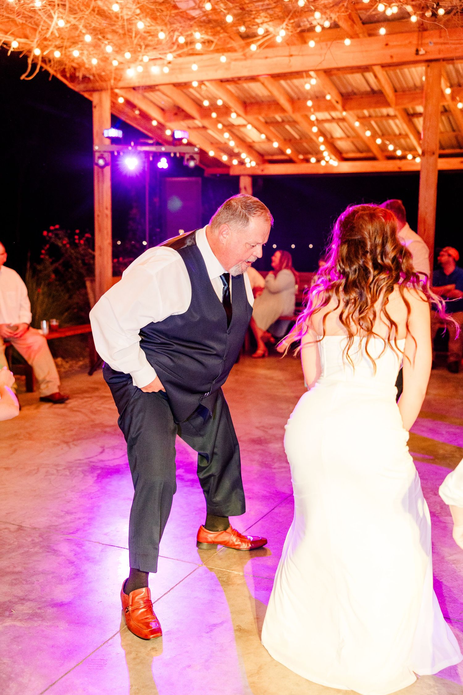 Bride and groom share a lively dance under string lights at an outdoor wedding reception with purple lighting.