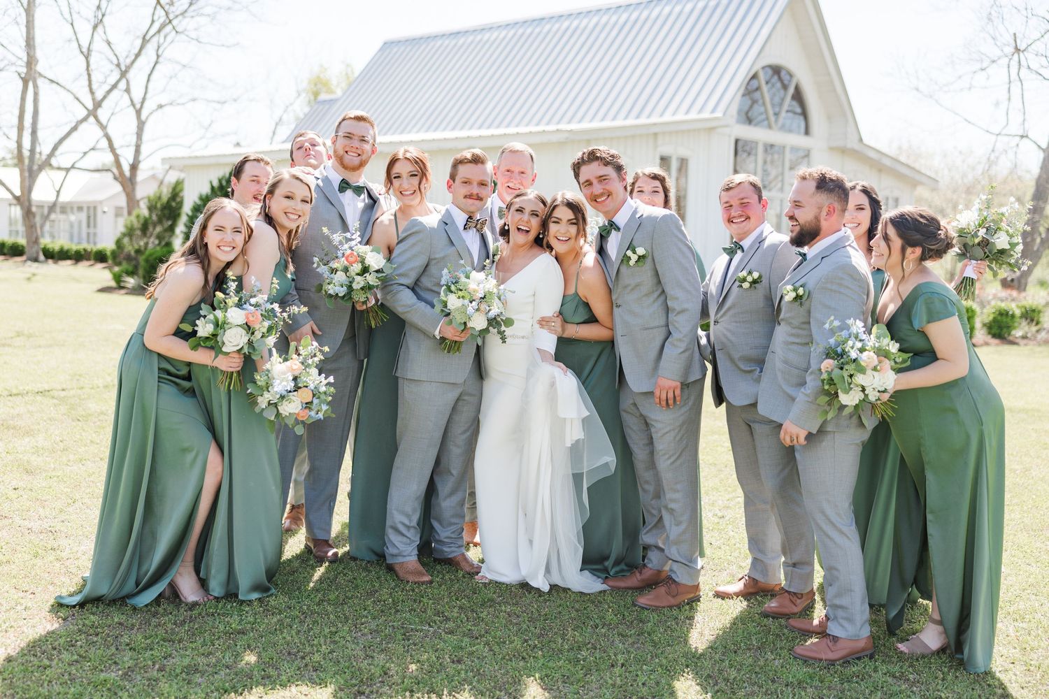 Wedding party in sage green dresses and grey suits posing outdoors on grass.