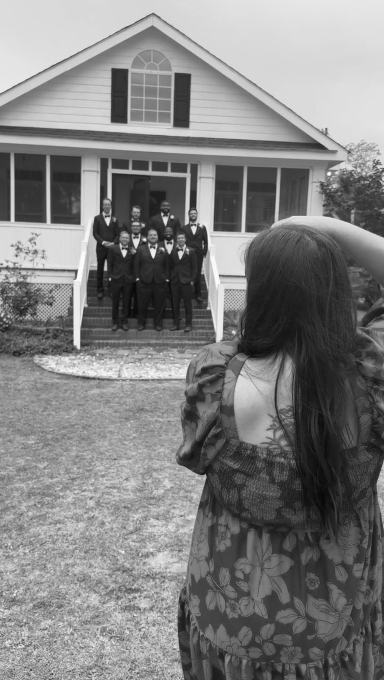 Woman in floral dress photographs groomsmen in suits posing on steps of white house in black and white photo.
