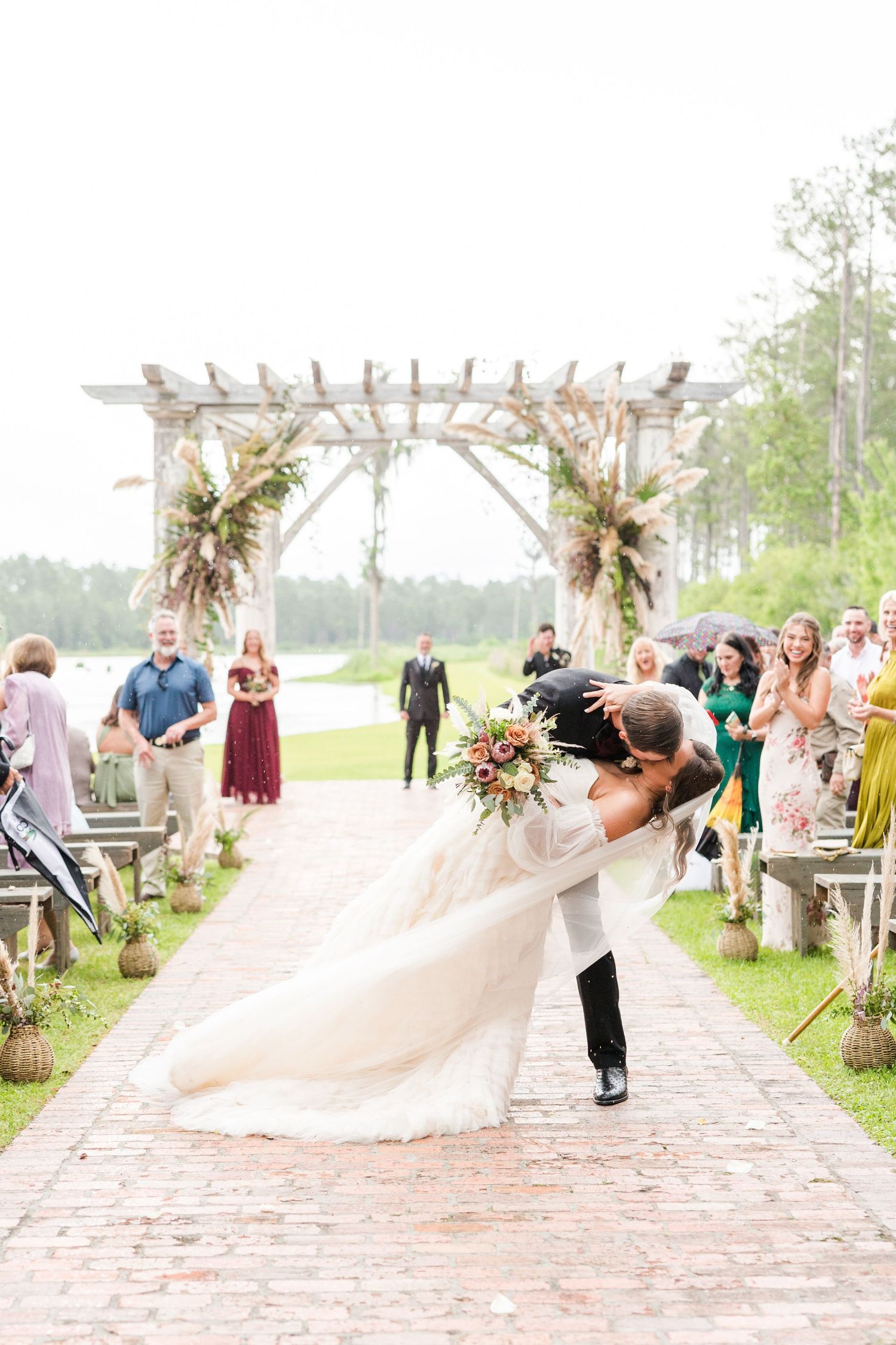 Romantic dip kiss under a floral-adorned wedding arch on a brick pathway.