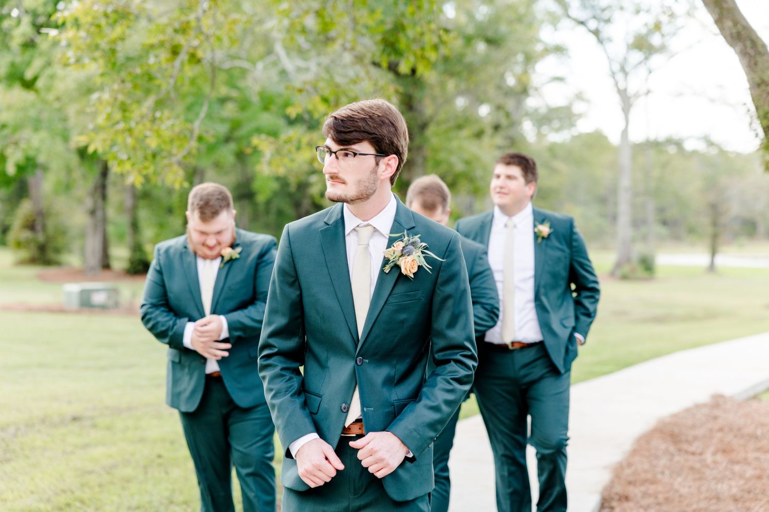 Groom in dark green suit with groomsmen in matching attire standing outdoors at a wedding ceremony.