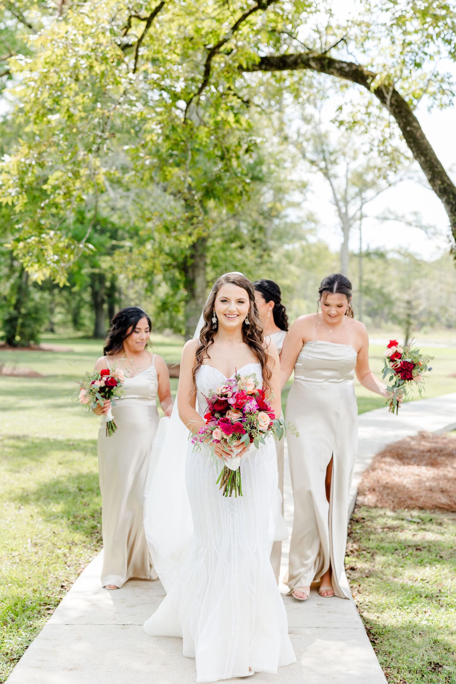 Bride in white gown walks with three bridesmaids in champagne dresses holding vibrant pink bouquets outdoors.