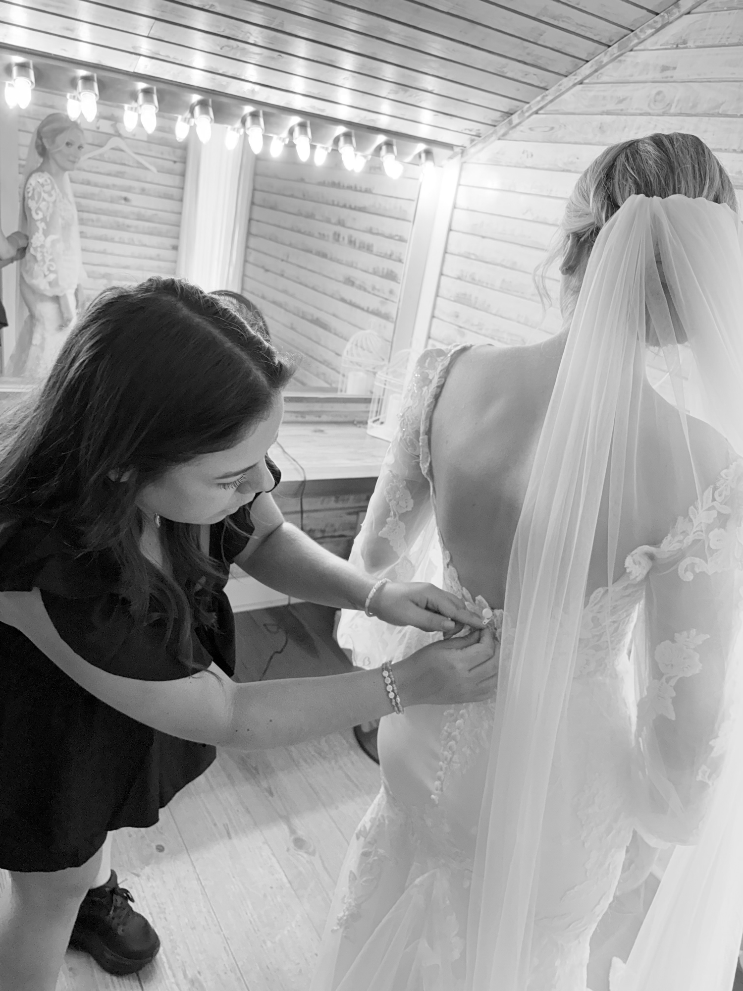 Black and white photo of a bridesmaid helping a bride button her wedding dress before the ceremony.