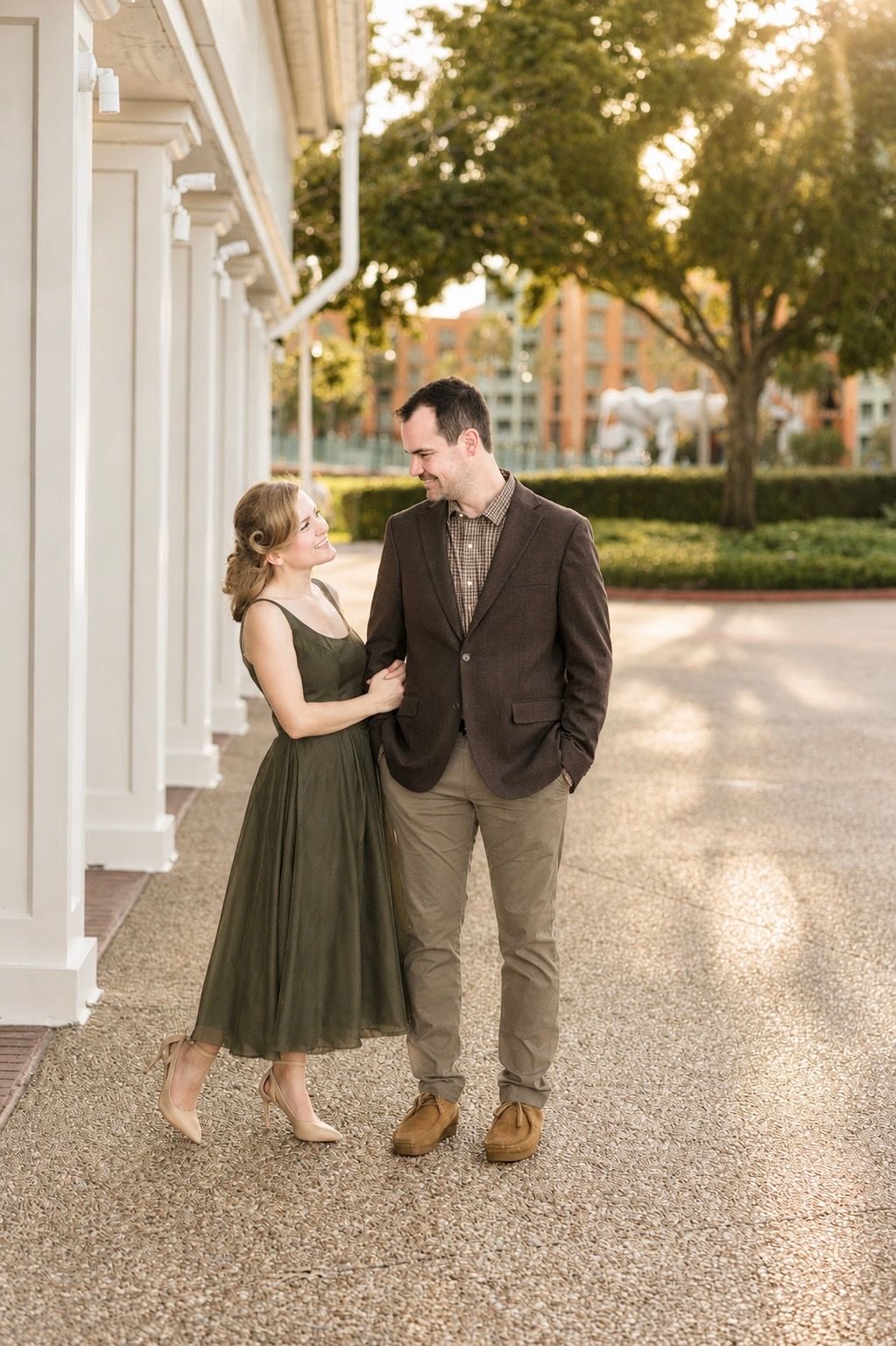 Couple in olive dress and brown suit smiling at each other outdoors near white columns at golden hour.